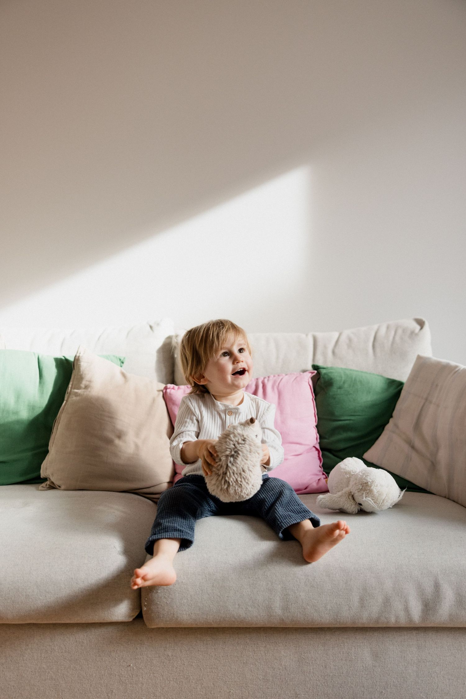 Toddler sitting on beige sofa with colorful pillows, holding stuffed animal toy, laughing in bright living room.