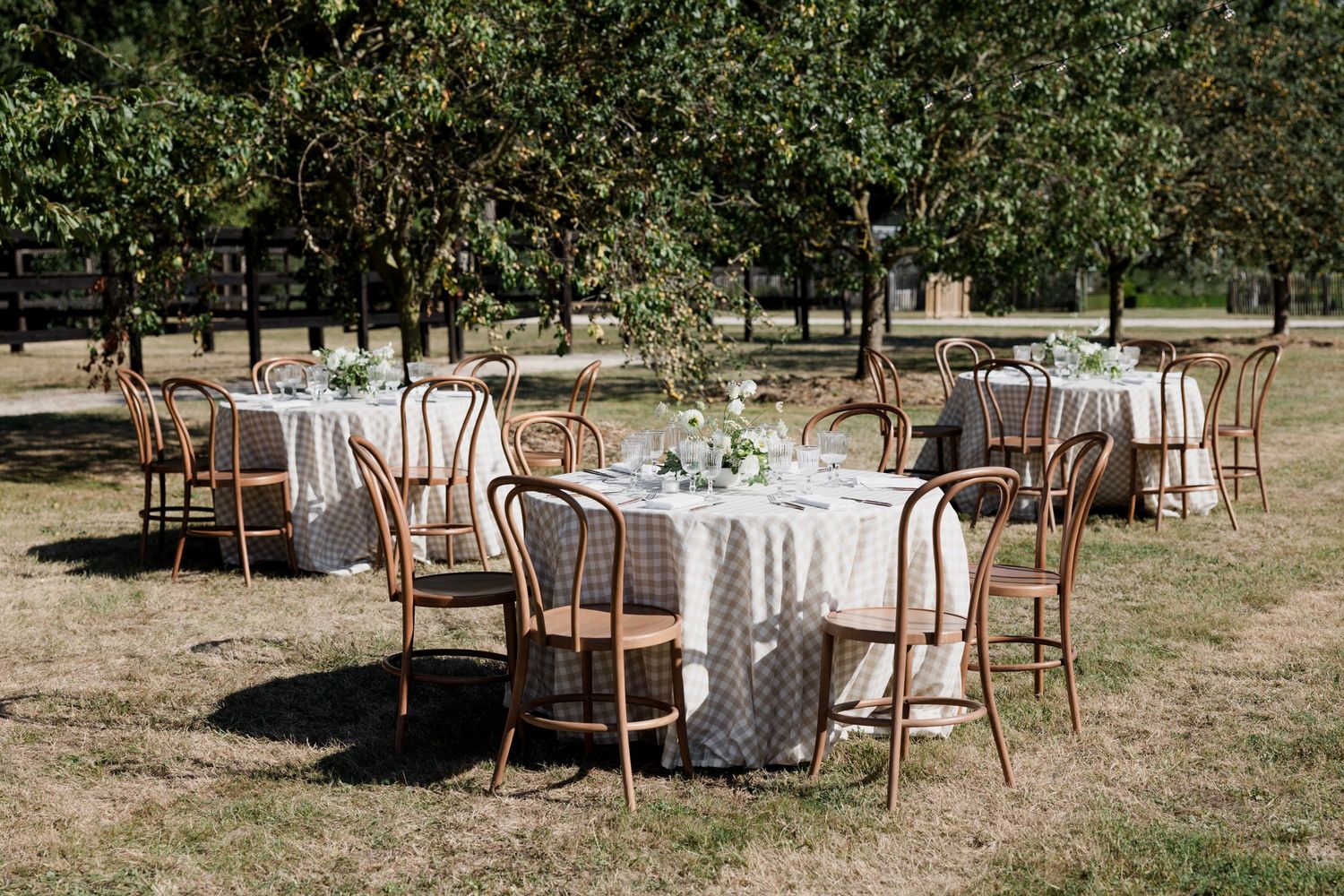 Elegant outdoor wedding reception setup with round tables, wooden chairs and lace tablecloths in a garden setting.