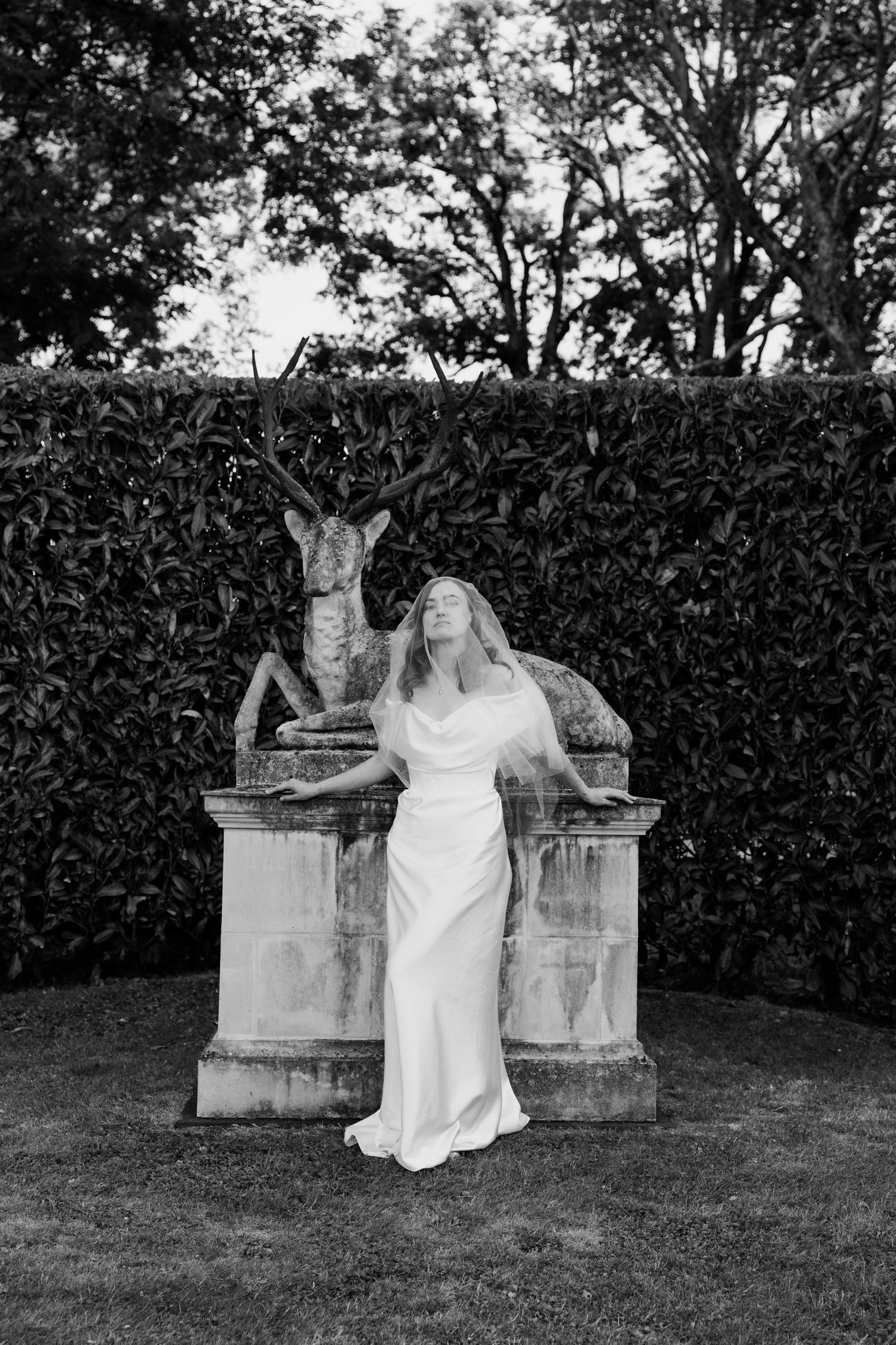 Black and white photo of a person in a white dress posing next to a decorative stone deer statue in a garden setting. Paris Tours wedding