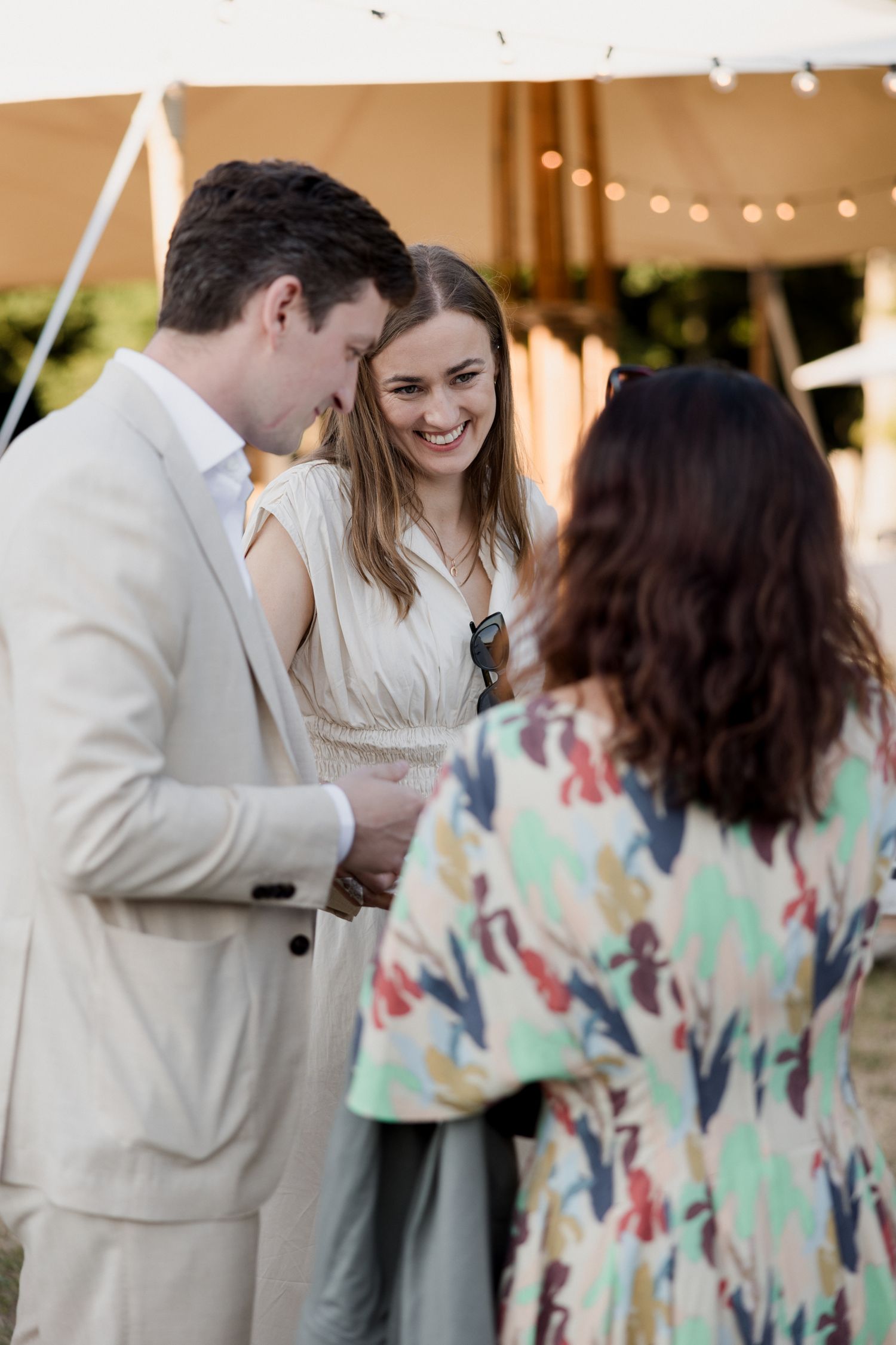 Wedding guests share joyful moments during an outdoor reception with string lights and floral dresses.