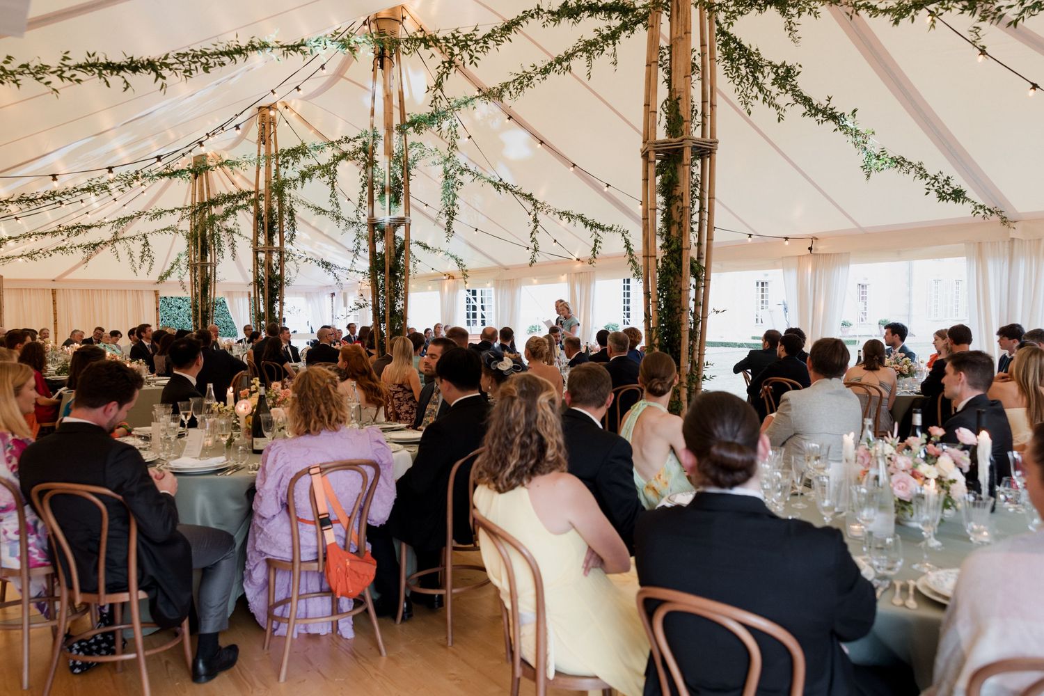 Wedding guests dine under an elegant white tent decorated with greenery garlands and string lights at a reception.