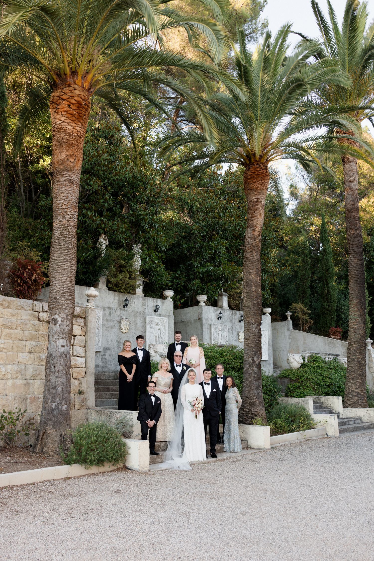 Wedding party poses for formal photos along a stone wall with palm trees in an elegant outdoor garden setting. chateau saint georges