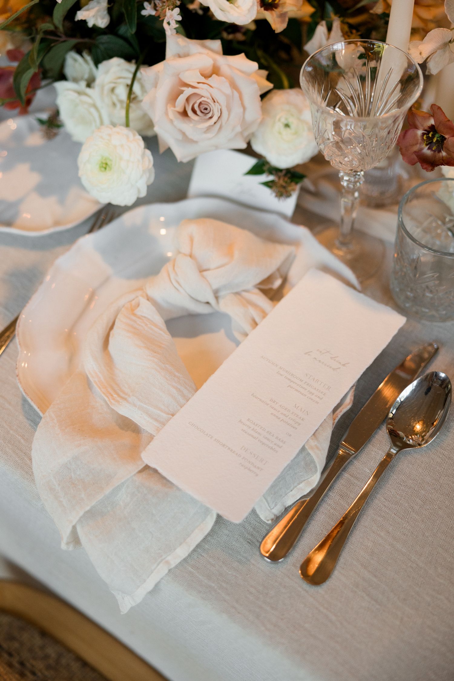 Elegant wedding table settings with blush pink menus, gold flatware, and soft floral centerpieces on neutral linens.