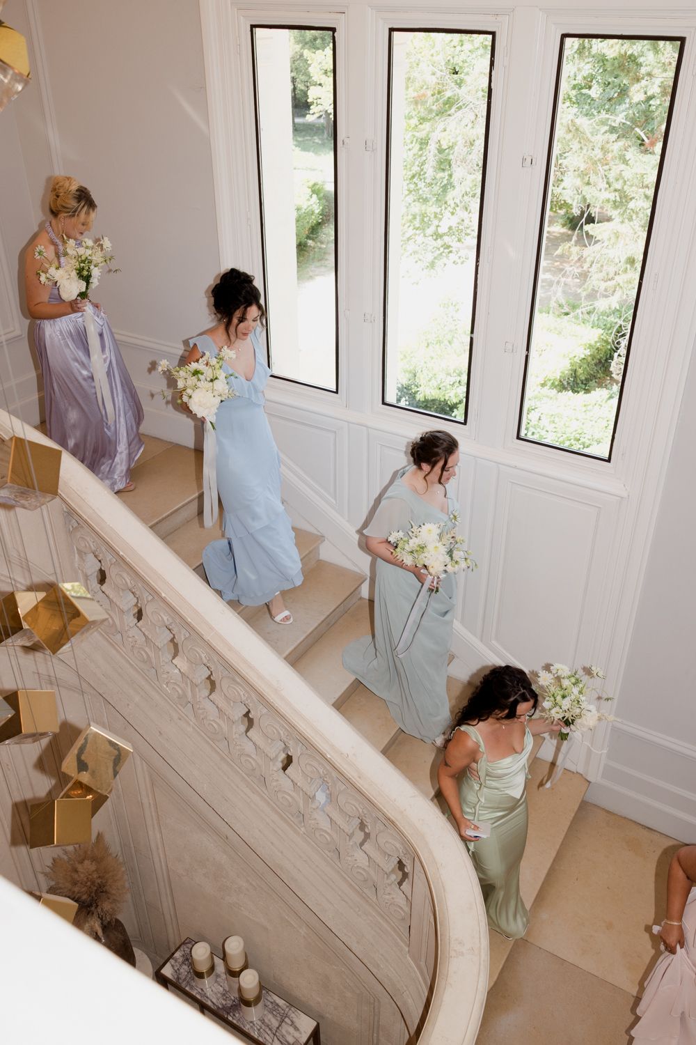 Bridesmaids in pastel dresses carrying white bouquets descend an elegant curved staircase with tall windows. Photographe mariage