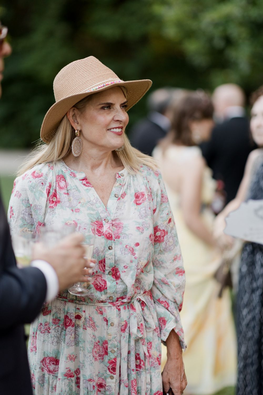 A person in a floral summer dress with puff sleeves and a beige hat smiles while attending an outdoor event.