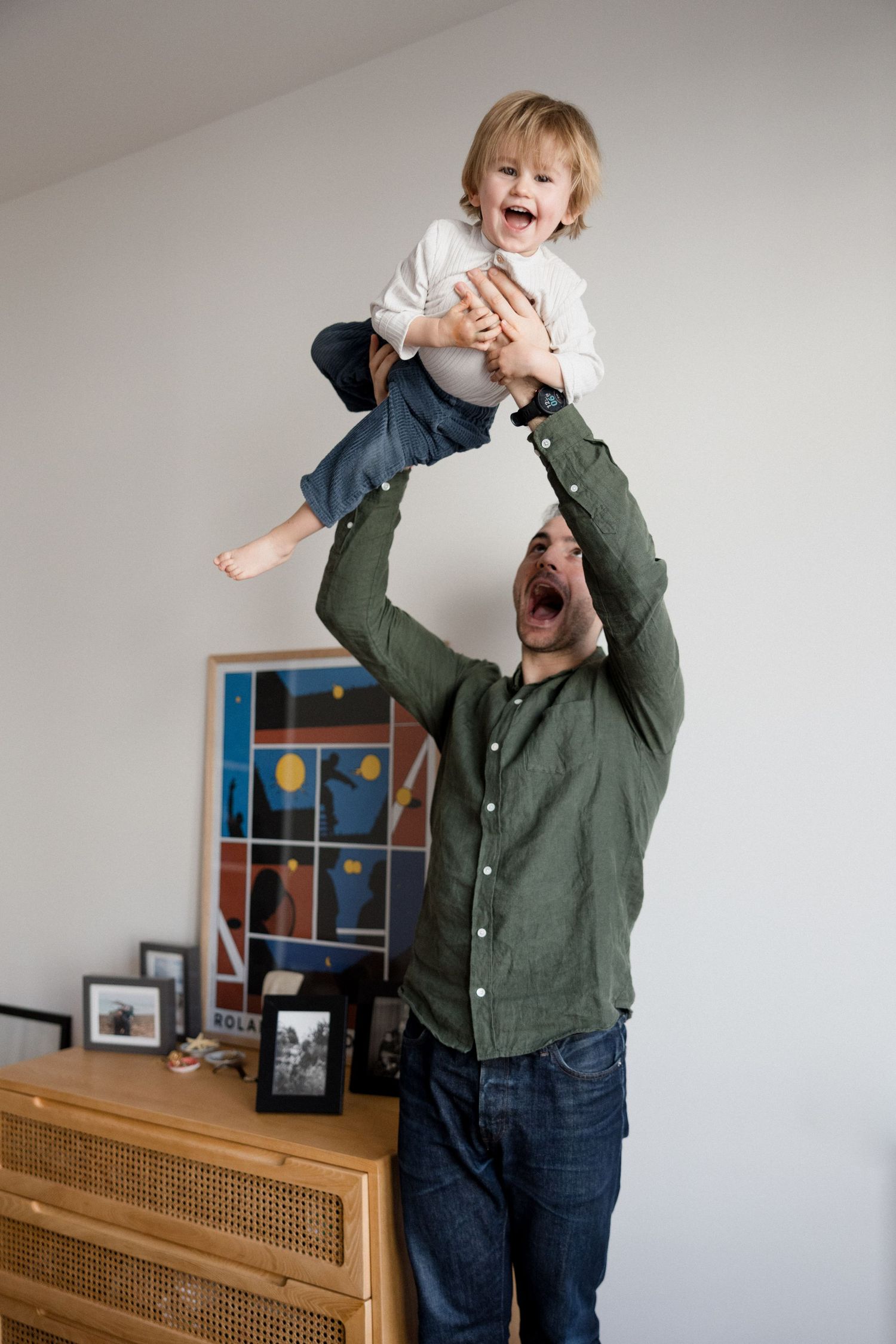 Father joyfully lifting laughing toddler above his head in a bright living room with artwork on the wall.