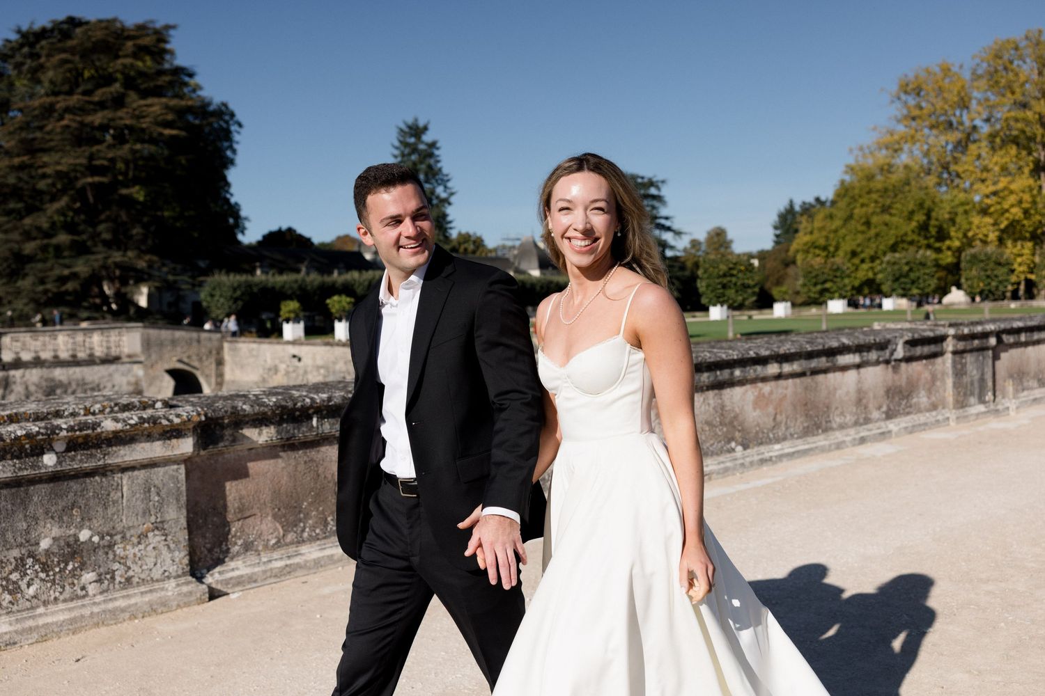 Bride and groom holding hands smiling at camera in elegant wedding attire outdoors in a sunny garden.