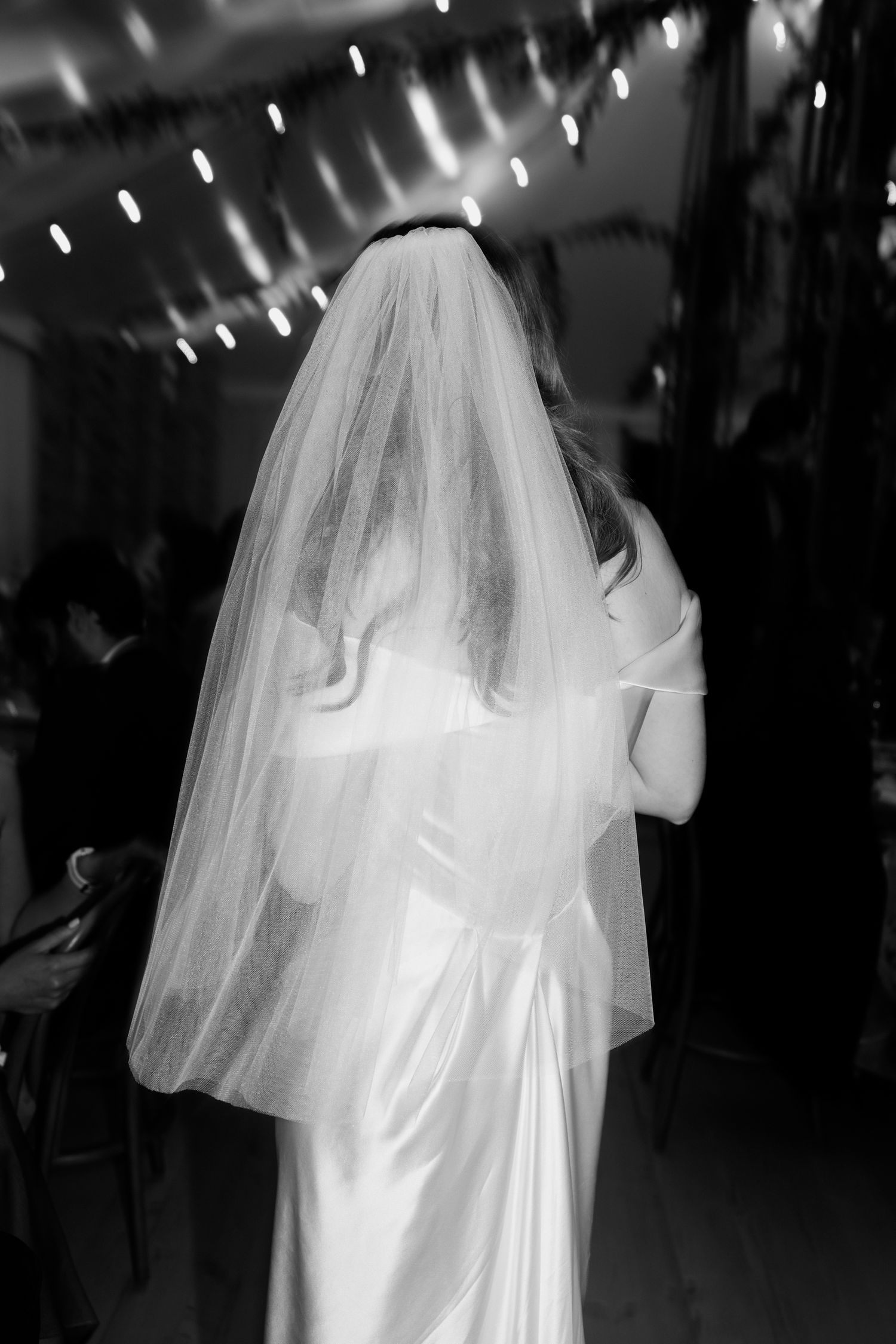 A bride in a white wedding dress and veil is shown from behind during a dance at a dimly lit reception venue.