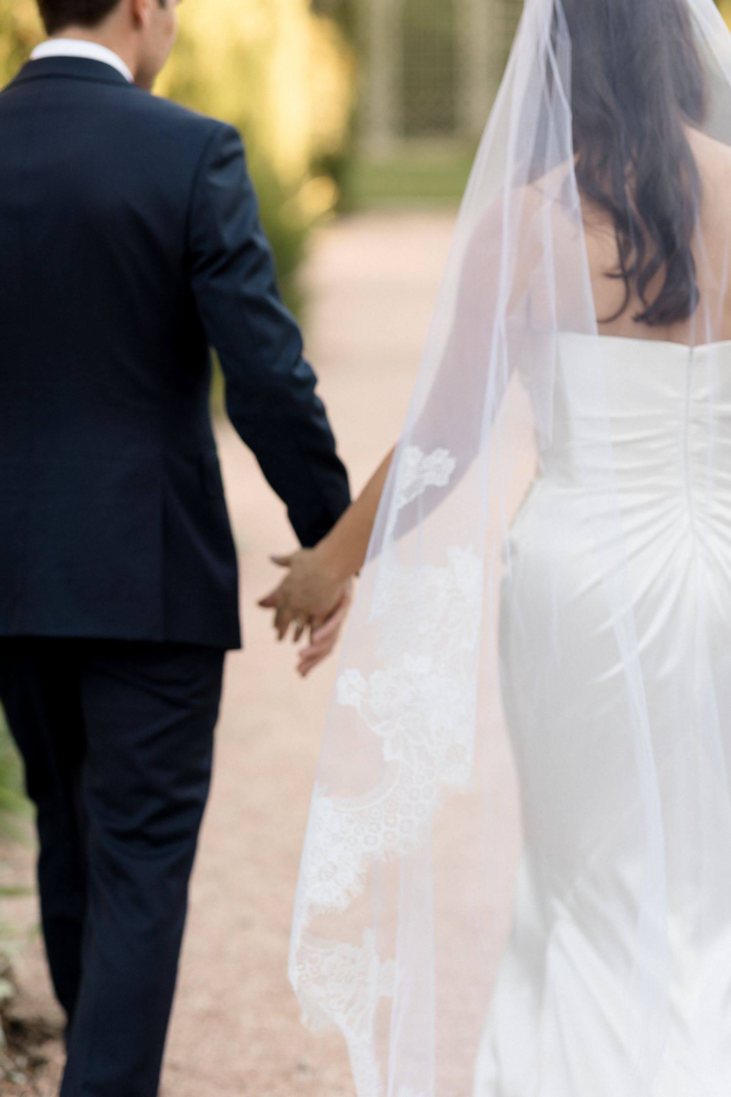 Close-up view of a bride and groom walking hand in hand, showing the back of her white dress and flowing veil.