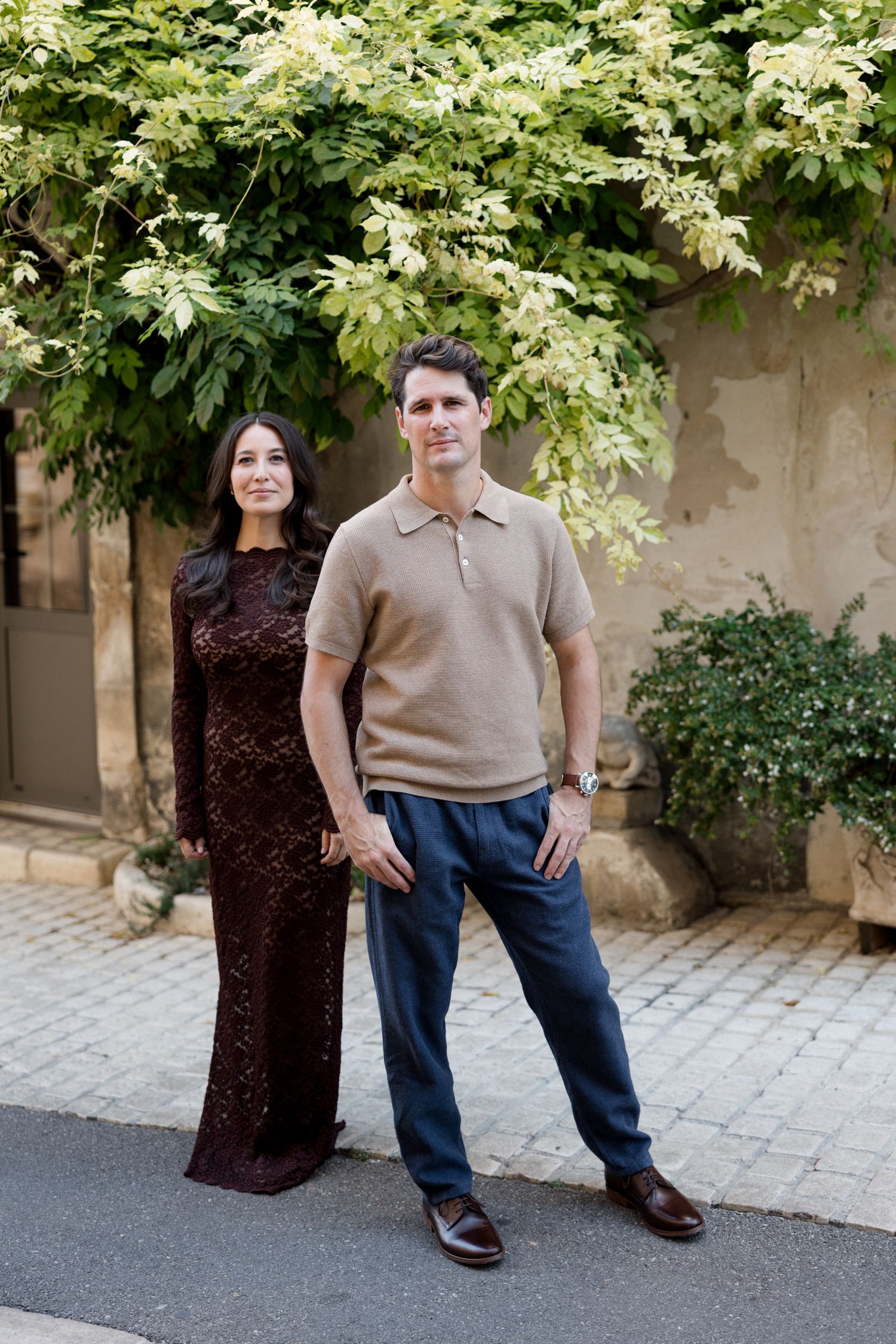 Two people stand together on a stone paved path next to a tree with green foliage in the background. Engagement session France