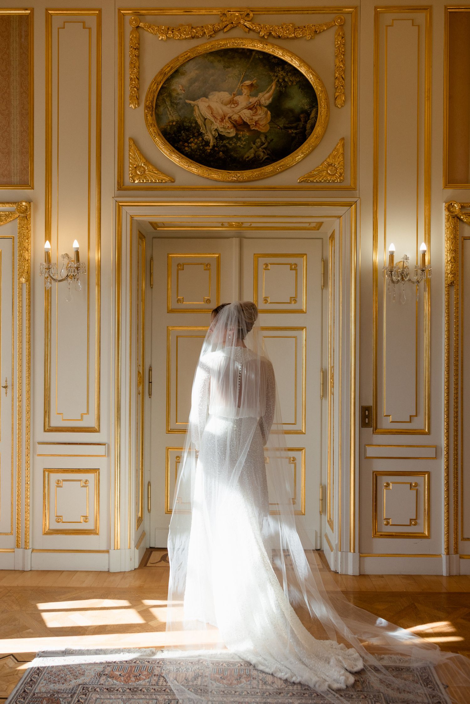 Elegant wedding dress with long veil photographed in an ornate gold-trimmed room with sunlight streaming through windows.