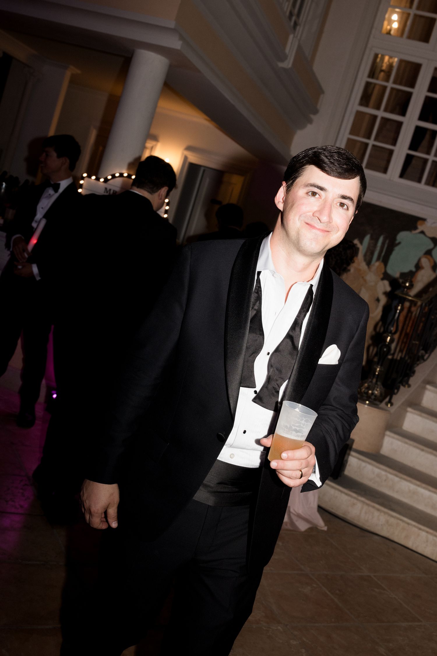 A person in formal black tuxedo and white shirt poses at an elegant indoor event venue.