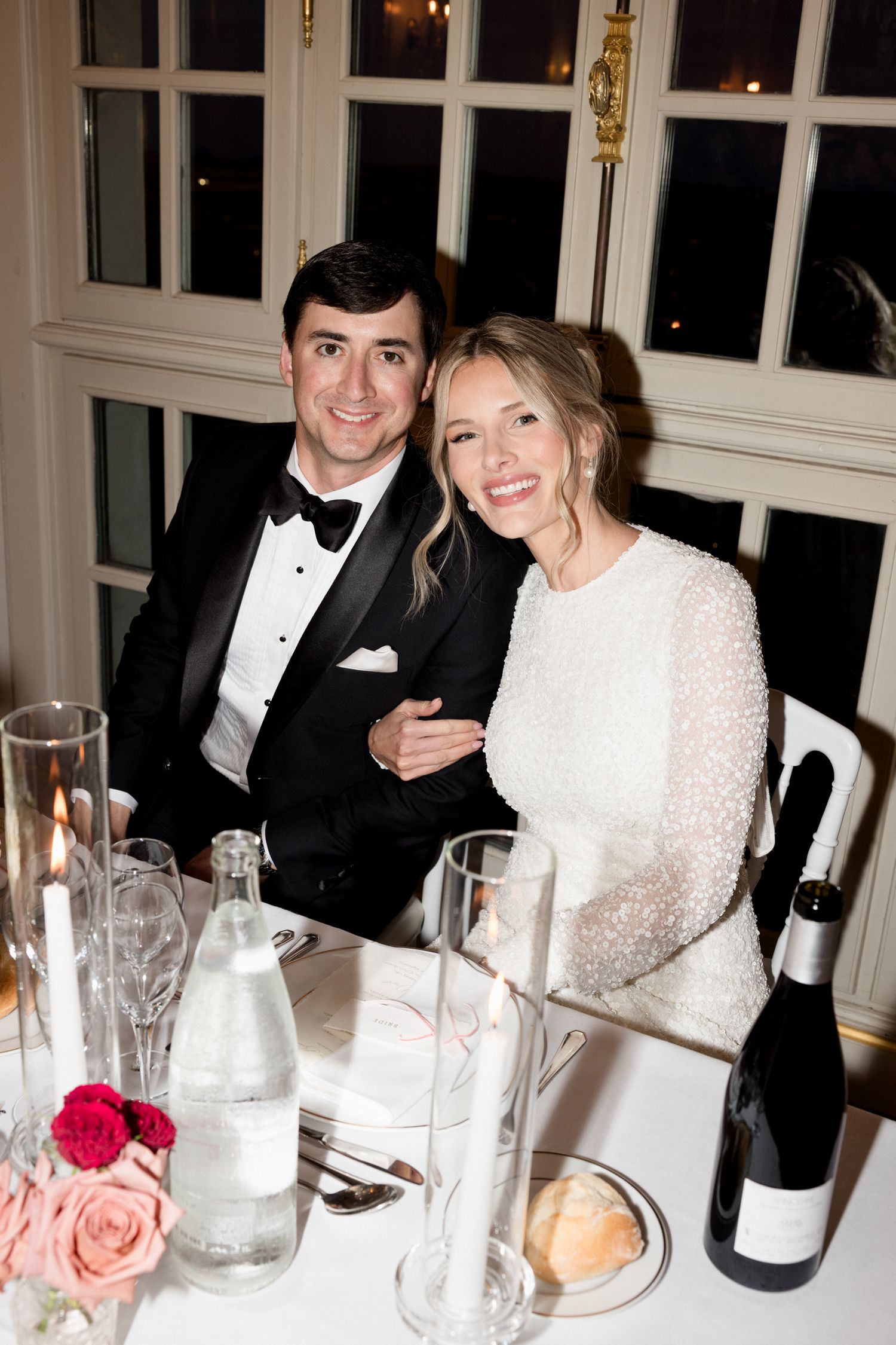 Elegant dinner party setting with people in formal attire seated at a table with wine glasses and floral centerpieces.