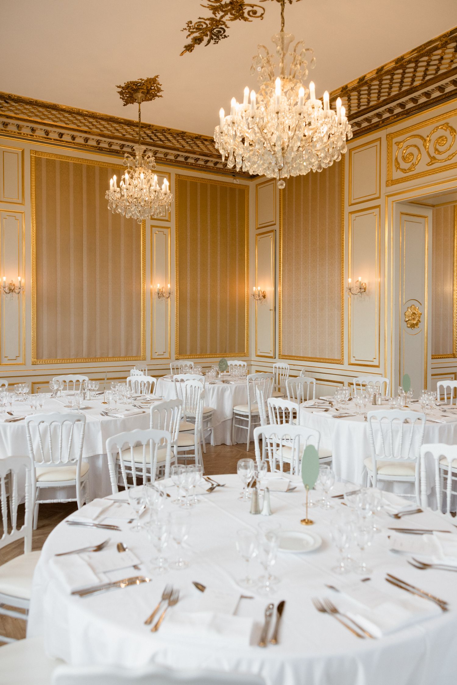 Elegant wedding reception venue with crystal chandeliers, white chairs, and formal table settings in a grand ballroom.