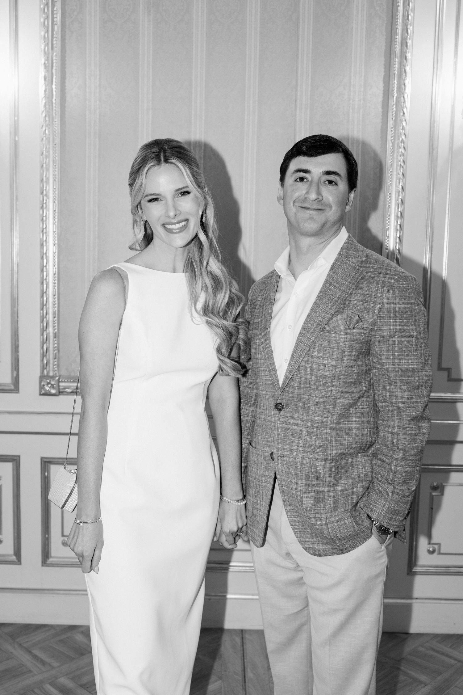 A black and white photo of a couple posing together, with her in a white dress and him wearing a plaid blazer.