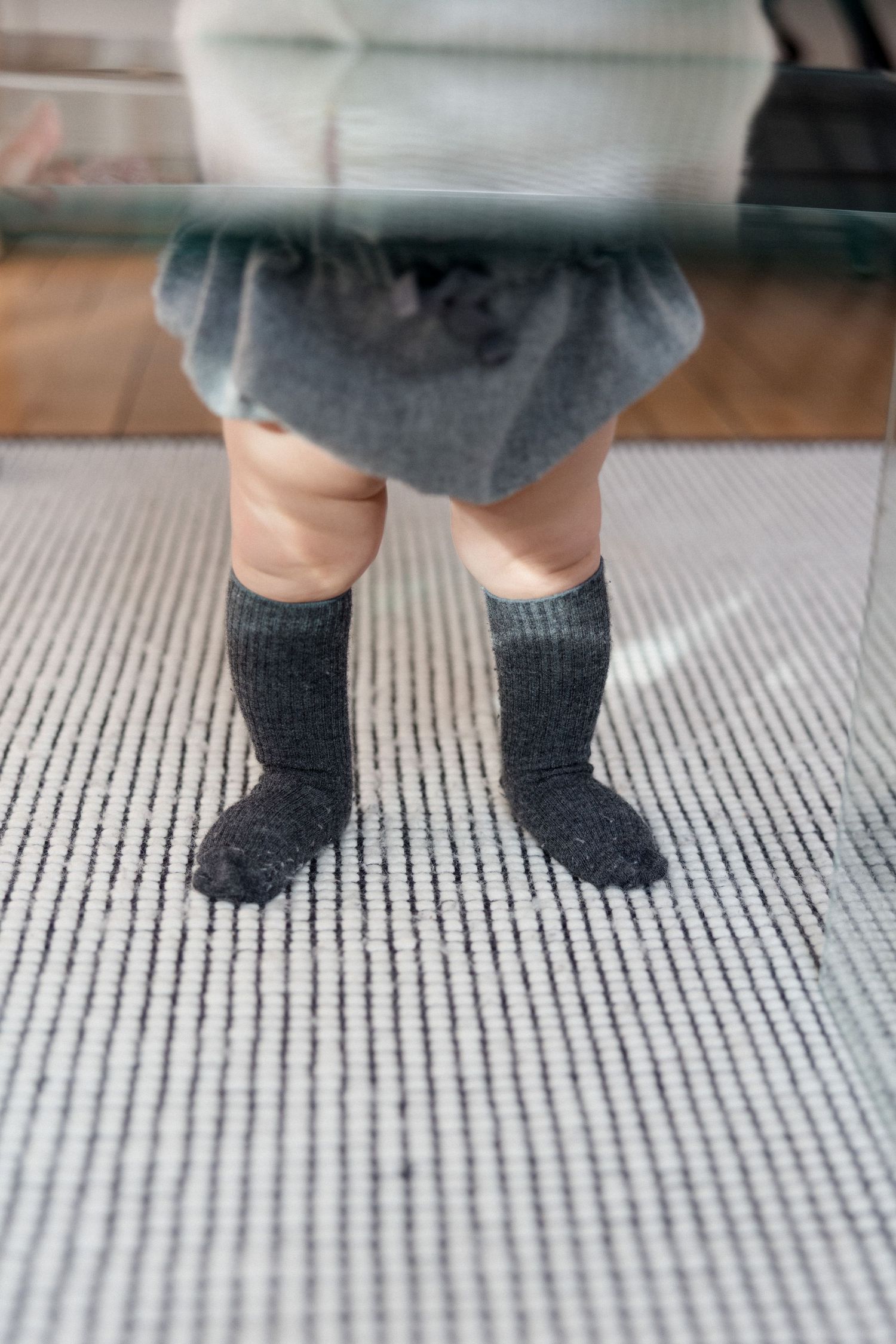 Toddler legs in dark gray knee-high socks and gray skirt standing on a striped carpet indoors.