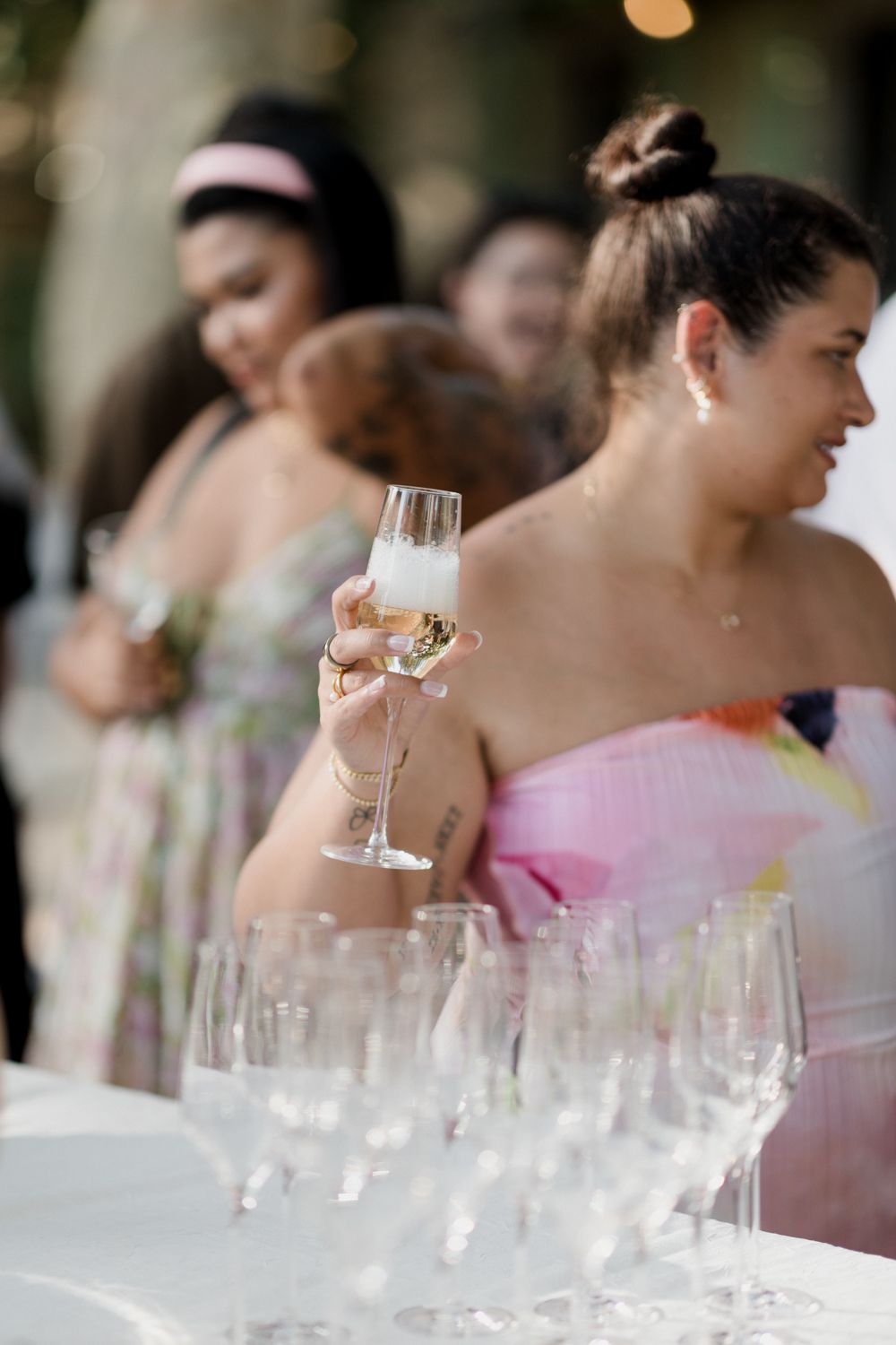 A person in a floral pink dress holds up a champagne glass at an elegant event with glassware on tables.