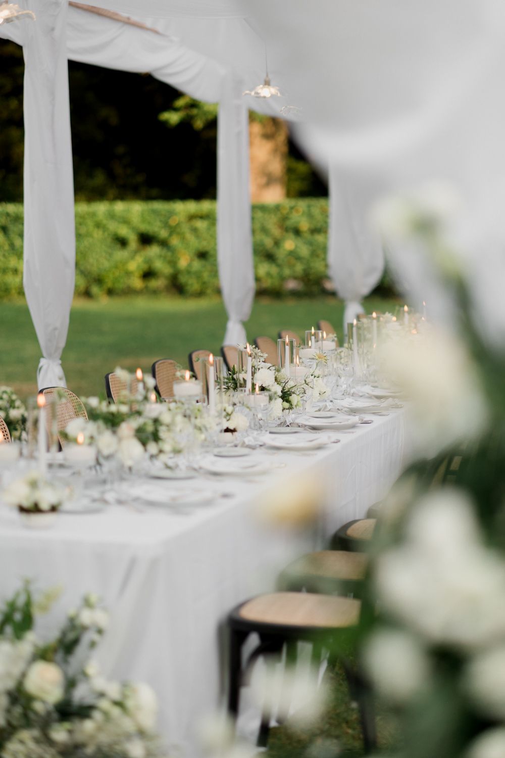 Elegant white wedding reception tables with floral centerpieces and draped fabric decorations in an outdoor garden setting. Horizon Wedding