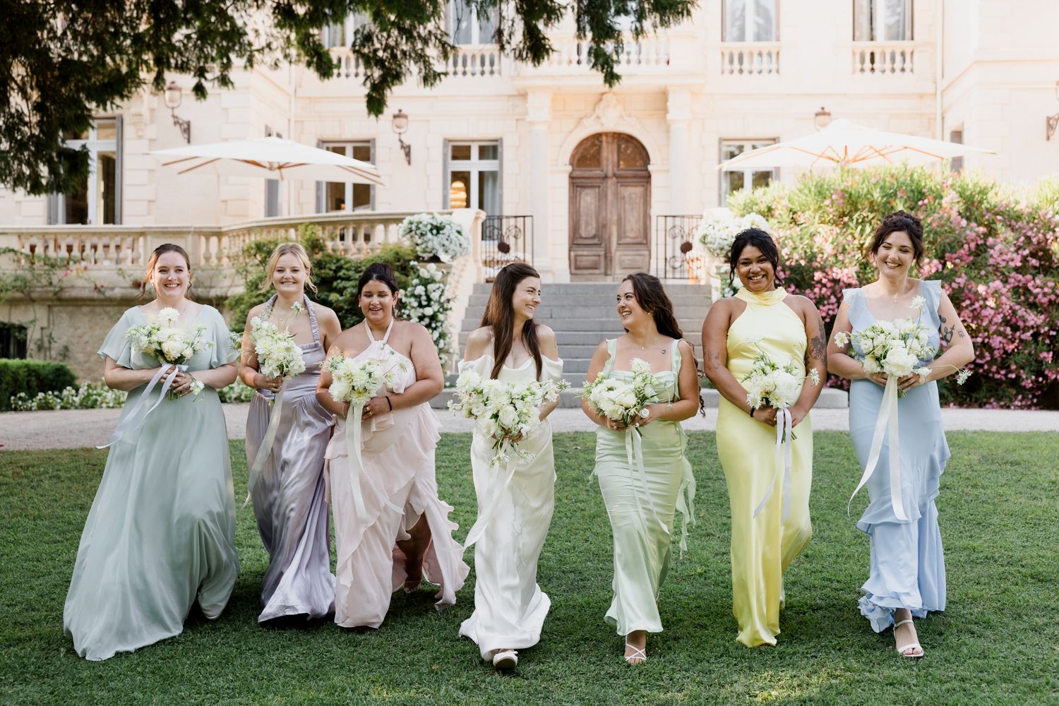 Delightful wedding party lineup of bridesmaids in pastel dresses skipping and twirling together on a sunny lawn. Château des 3 Fontaines