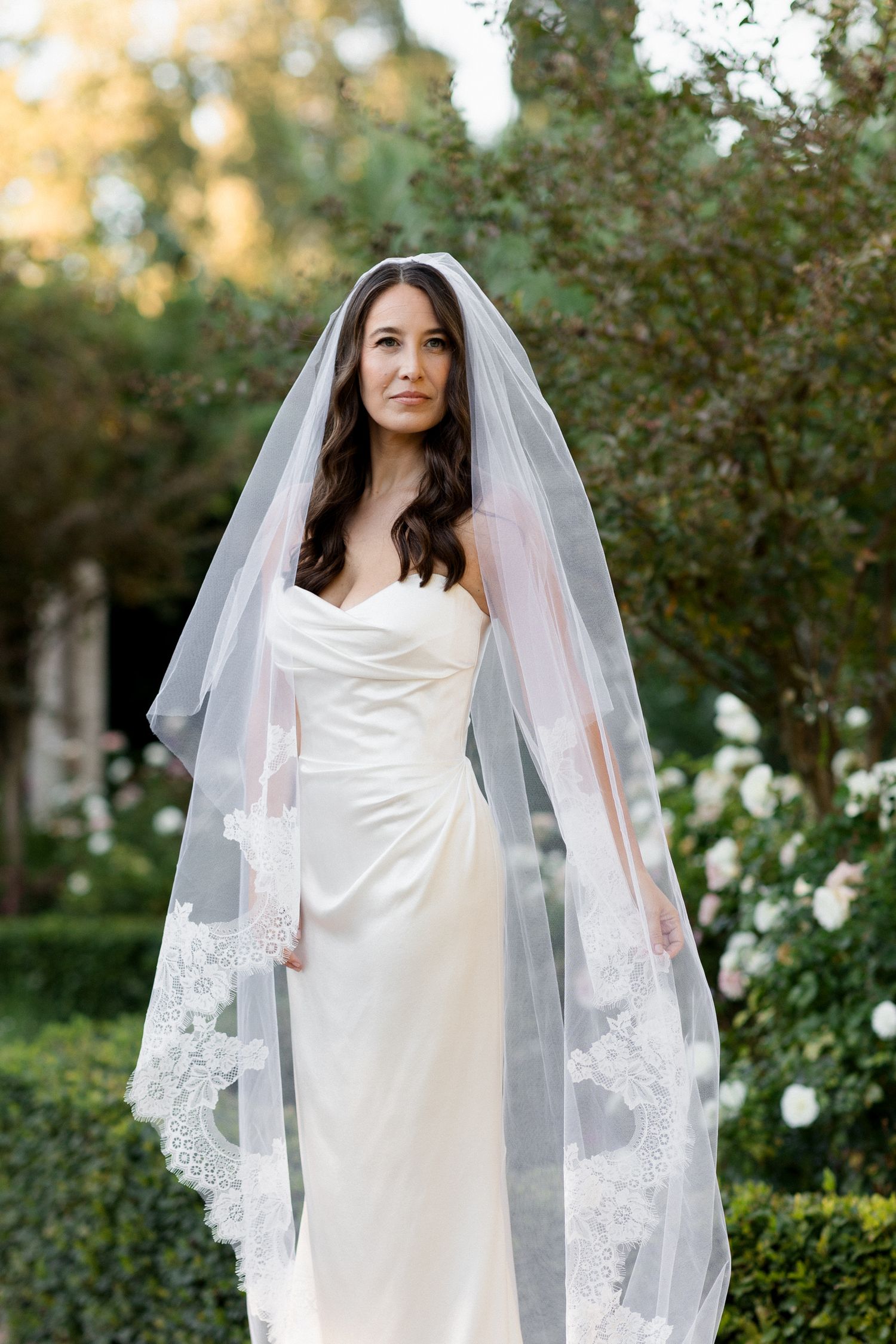 A bride in a classic white wedding dress and long veil poses elegantly in a garden setting with greenery and flowers.