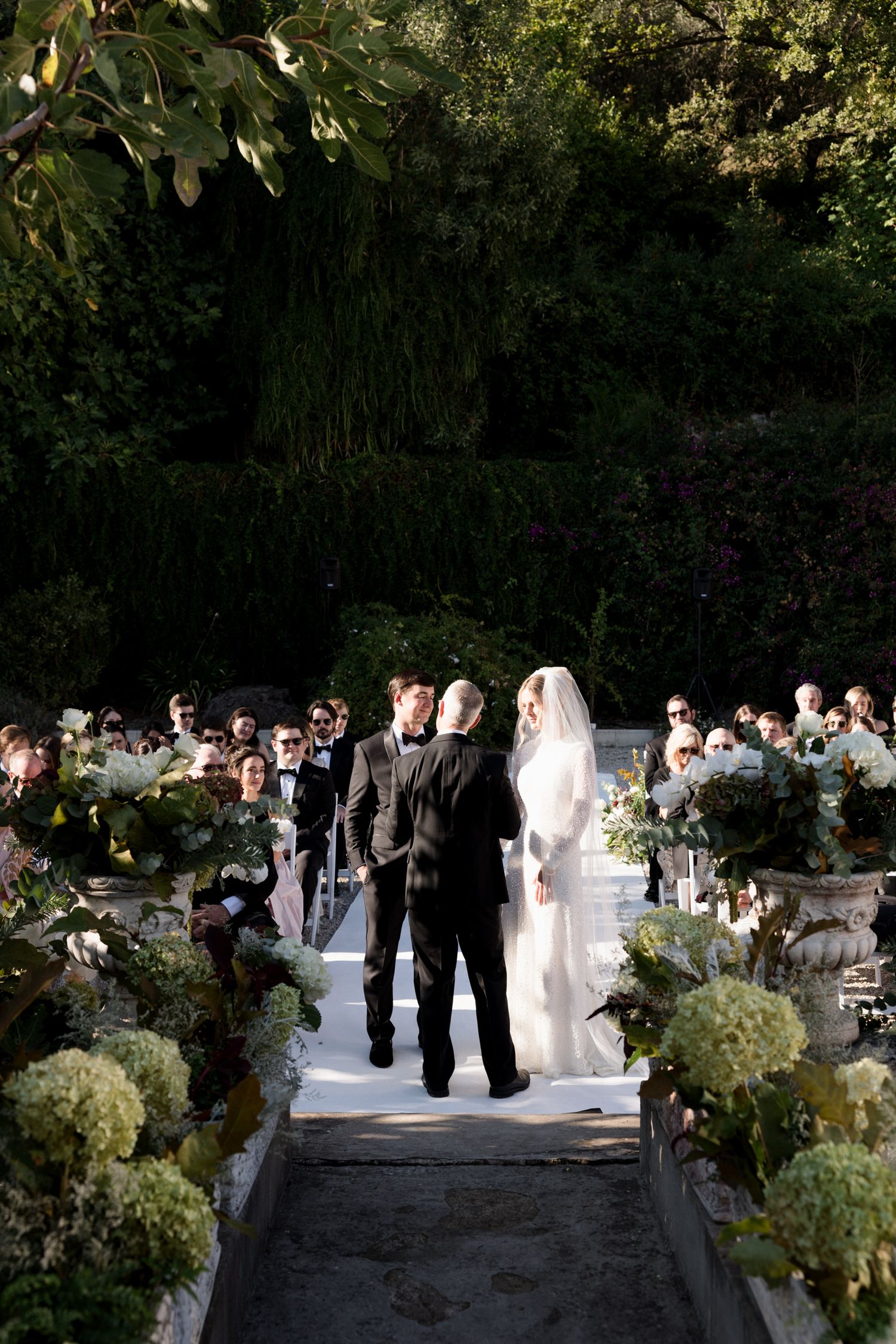 An elegant outdoor wedding ceremony in a garden setting with white floral arrangements lining the aisle.