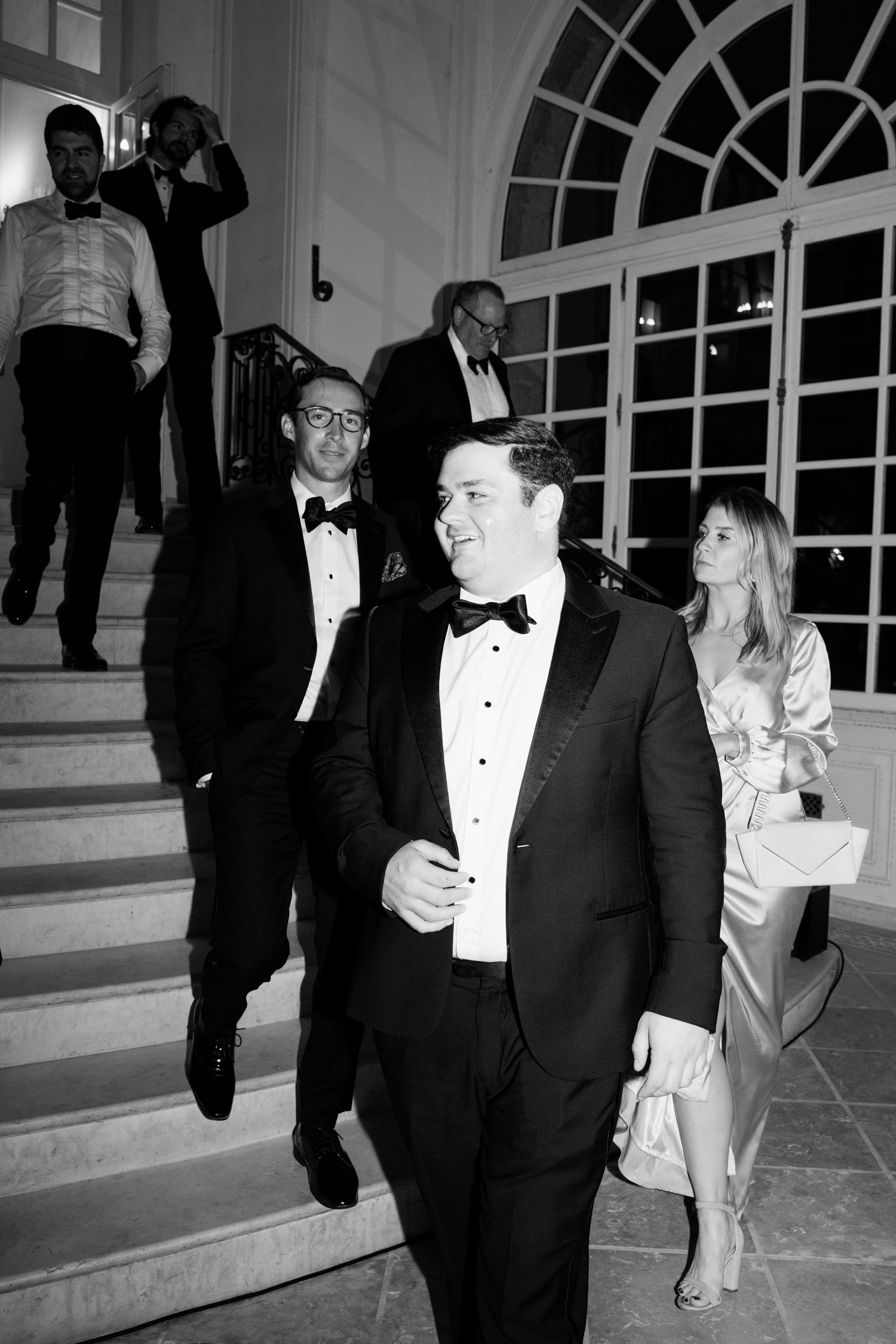 Group of people in formal black tie attire walking and socializing at an elegant venue with arched windows.