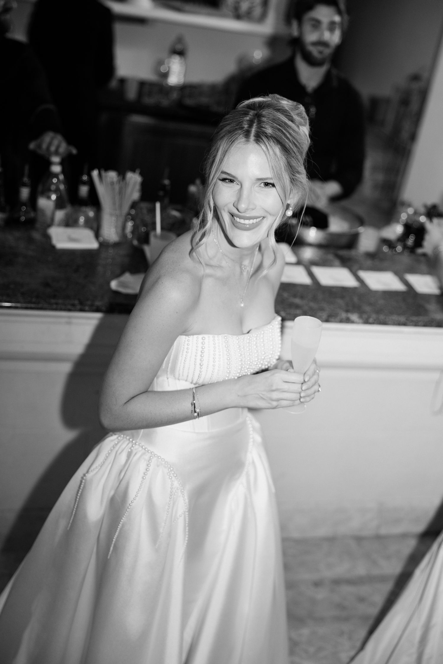A candid black and white photo of someone in a white strapless wedding dress smiling at a bar reception. Editorial wedding photographer fran