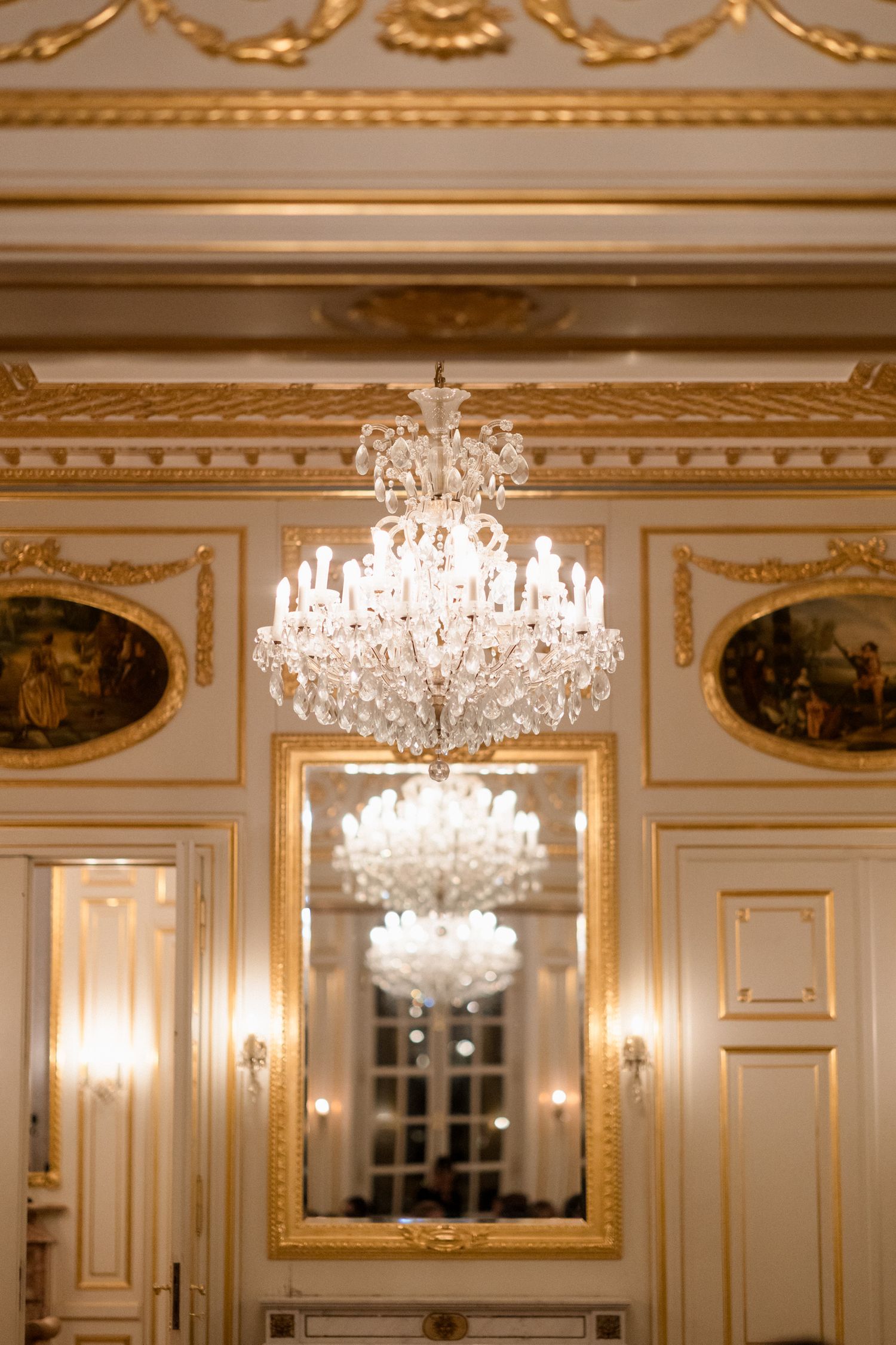 Elegant crystal chandelier hangs in ornate room with gold-trimmed walls, mirrors and classical oval paintings.