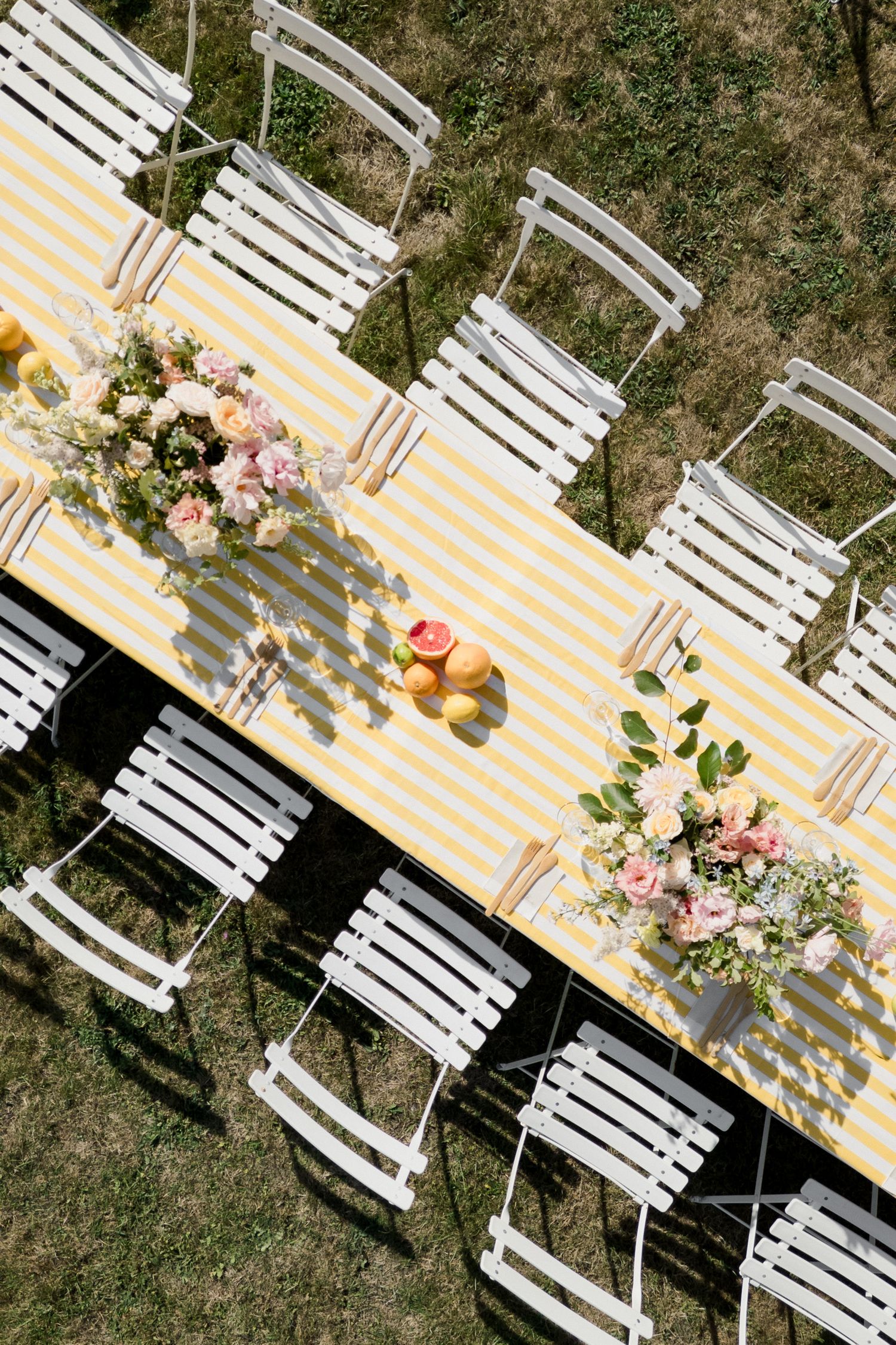 Overhead view of white garden chairs and a yellow striped tablecloth with floral centerpieces on a lawn.
