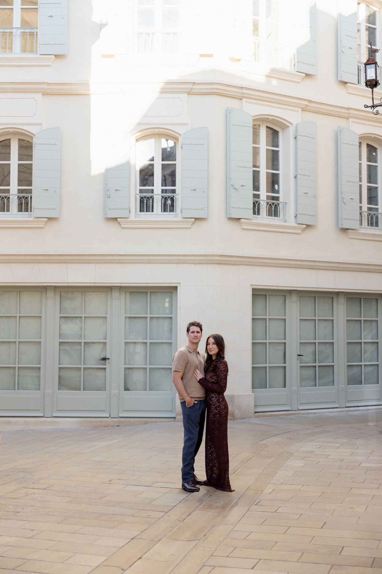 A couple stands together on a modern patio with white architectural walls and large windows in the background. Engagement session Paris