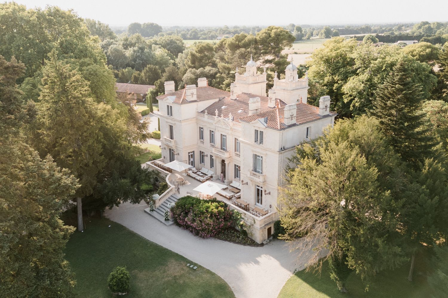 Elegant white French chateau with circular driveway surrounded by lush trees viewed from above on a sunny day. Château des 3 Fontaines