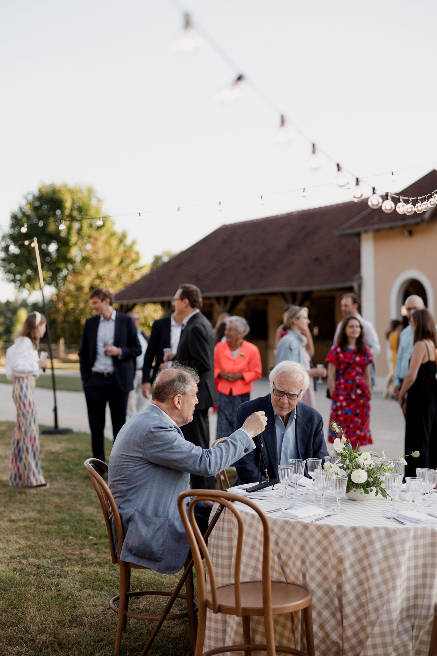 Outdoor wedding reception at sunset with guests seated at tables with checkered tablecloths near a rustic barn.