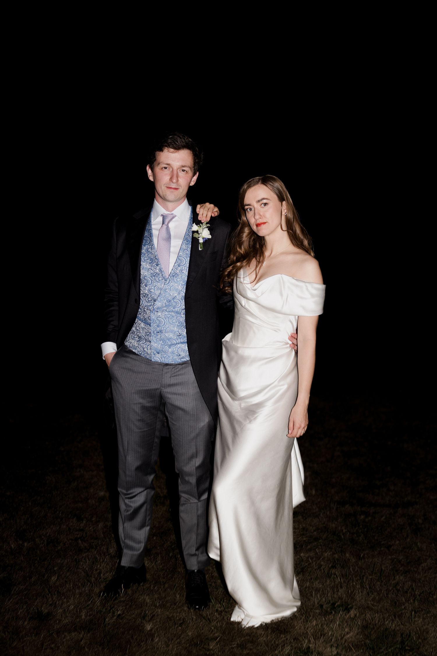 Wedding couple stands together at night in formal attire against dark background. Paris wedding photographer
