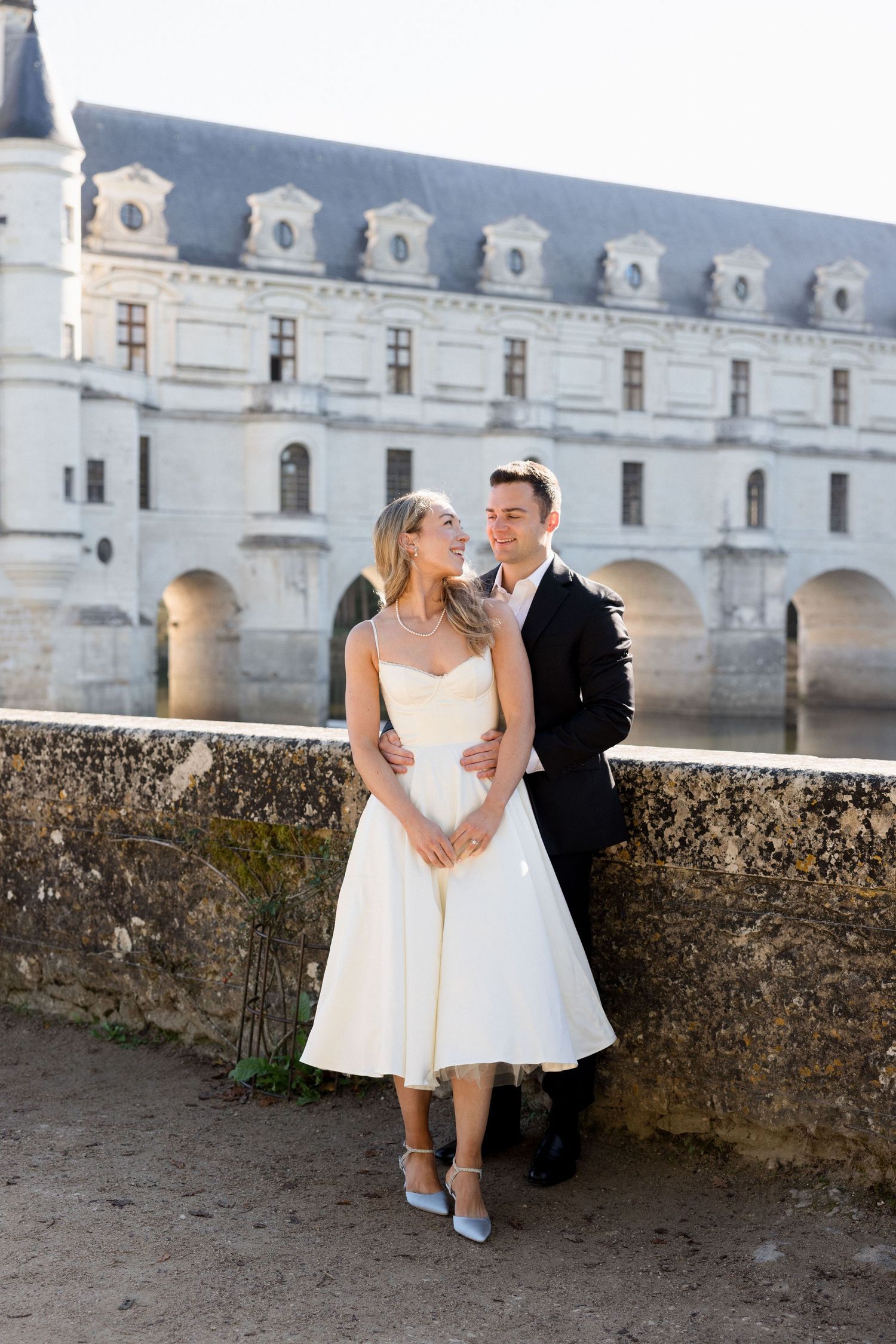 Bride and groom posing romantically in front of Chateau de Chenonceau, France, bride wearing tea-length white dress.