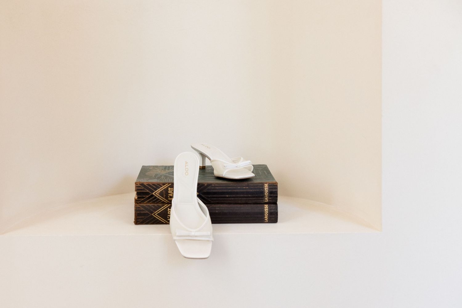 White strappy heeled sandals displayed on stacked dark books against a minimalist cream background.
