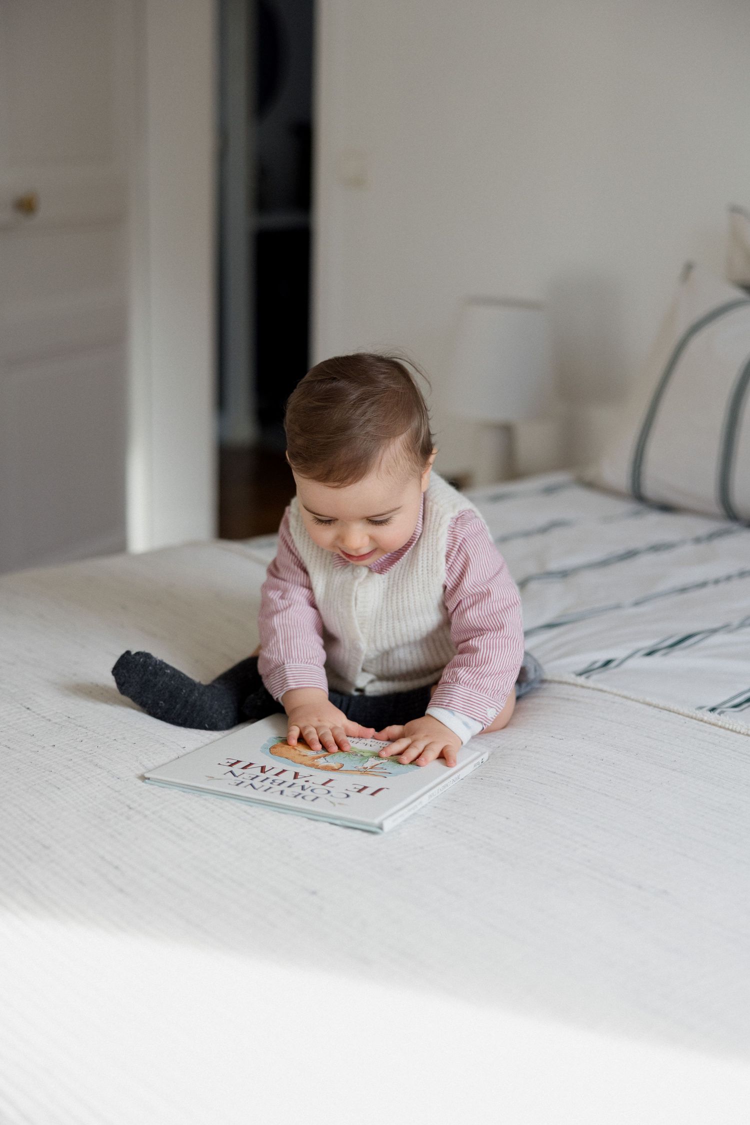 A cute baby sits on a white bed, smiling while looking at a colorful picture book.