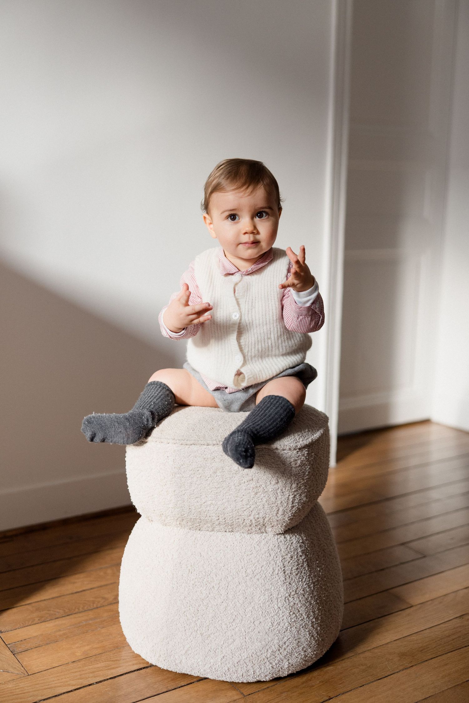 Adorable baby sitting on a white boucle pouf, wearing a cream cardigan and gray socks, reaching out tiny hands.