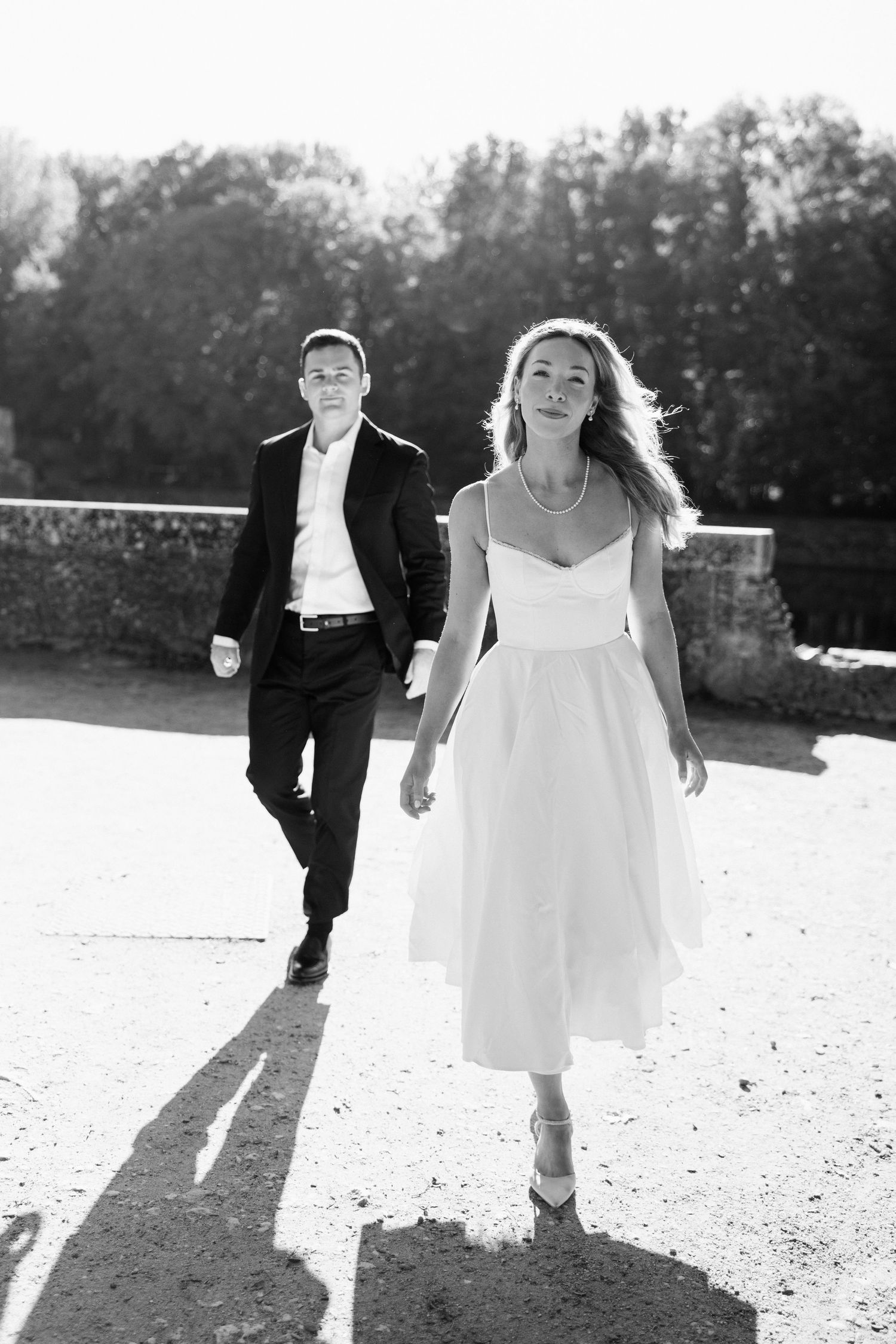 Black and white photo of bride and groom walking confidently toward camera on sunny stone bridge.