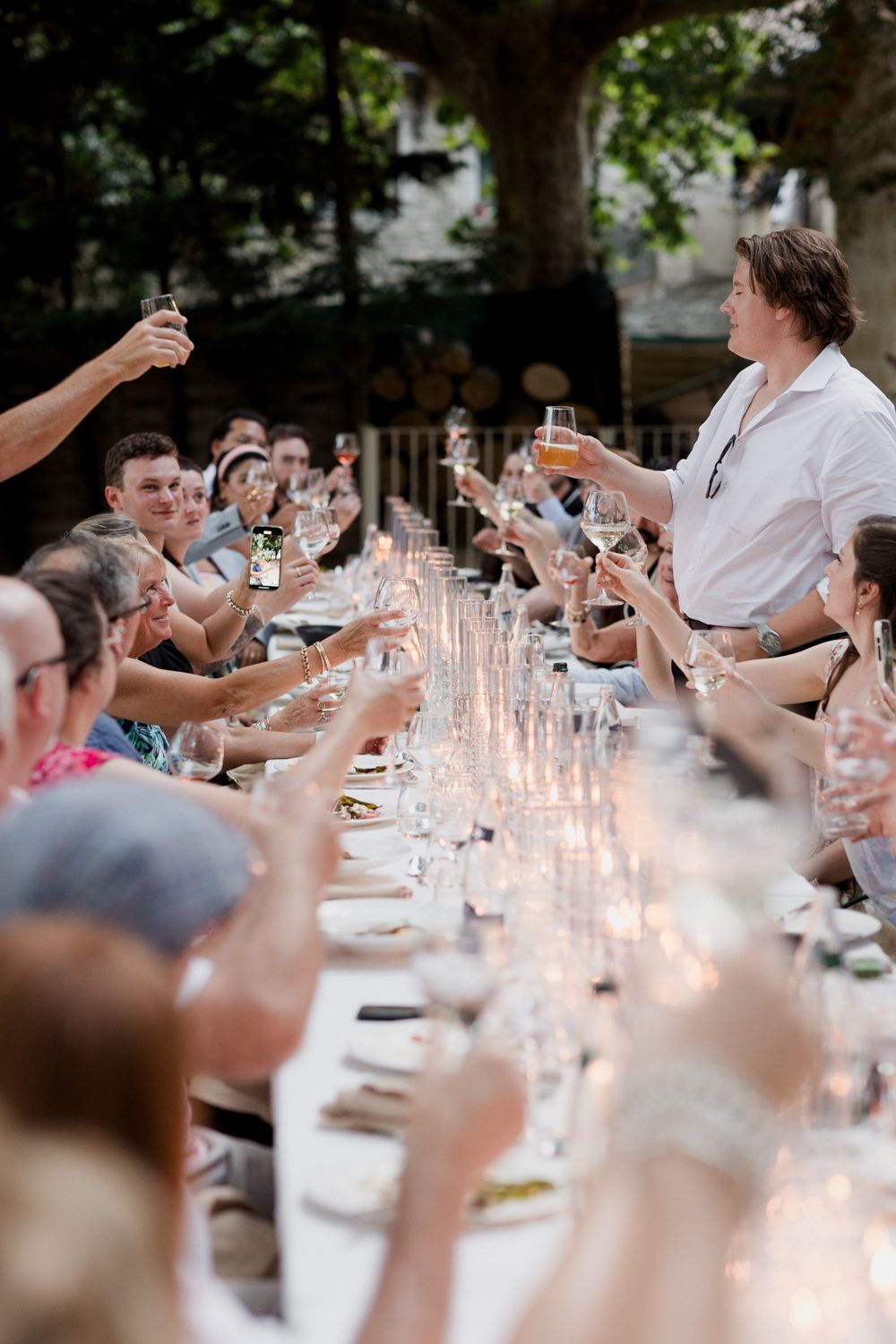 Guests raise wine glasses in a toast at an elegant outdoor dinner party with candlelit table settings at dusk.