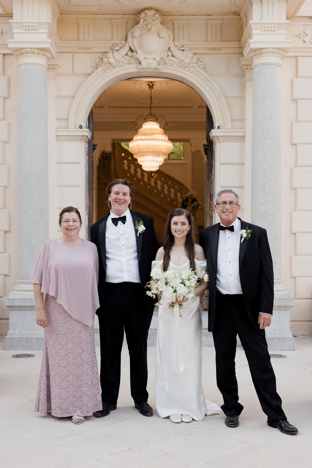 Wedding party poses in an elegant white marble hallway with classical columns and warm lighting.
