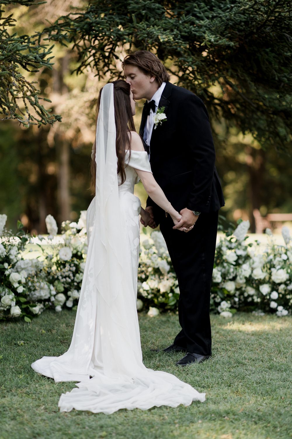 A beautiful outdoor wedding moment captured in a lush garden with white flowers in the background.