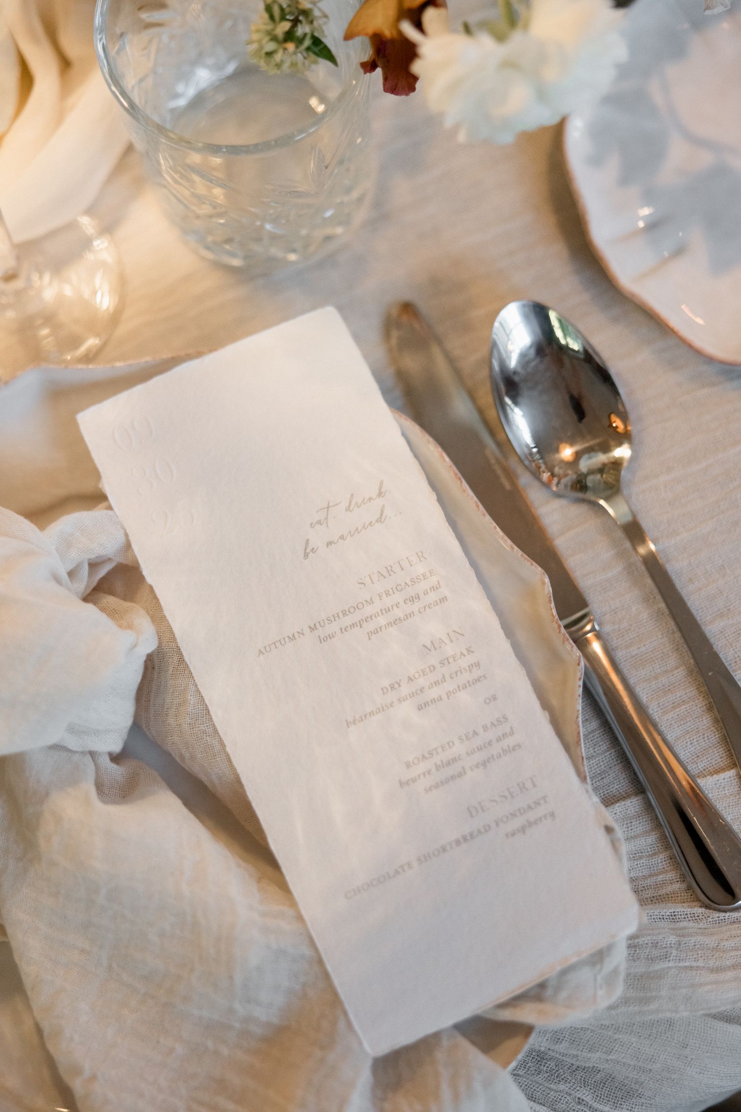 Elegant wedding menu card and silver spoon placed on white linen tablecloth at formal dinner setting.
