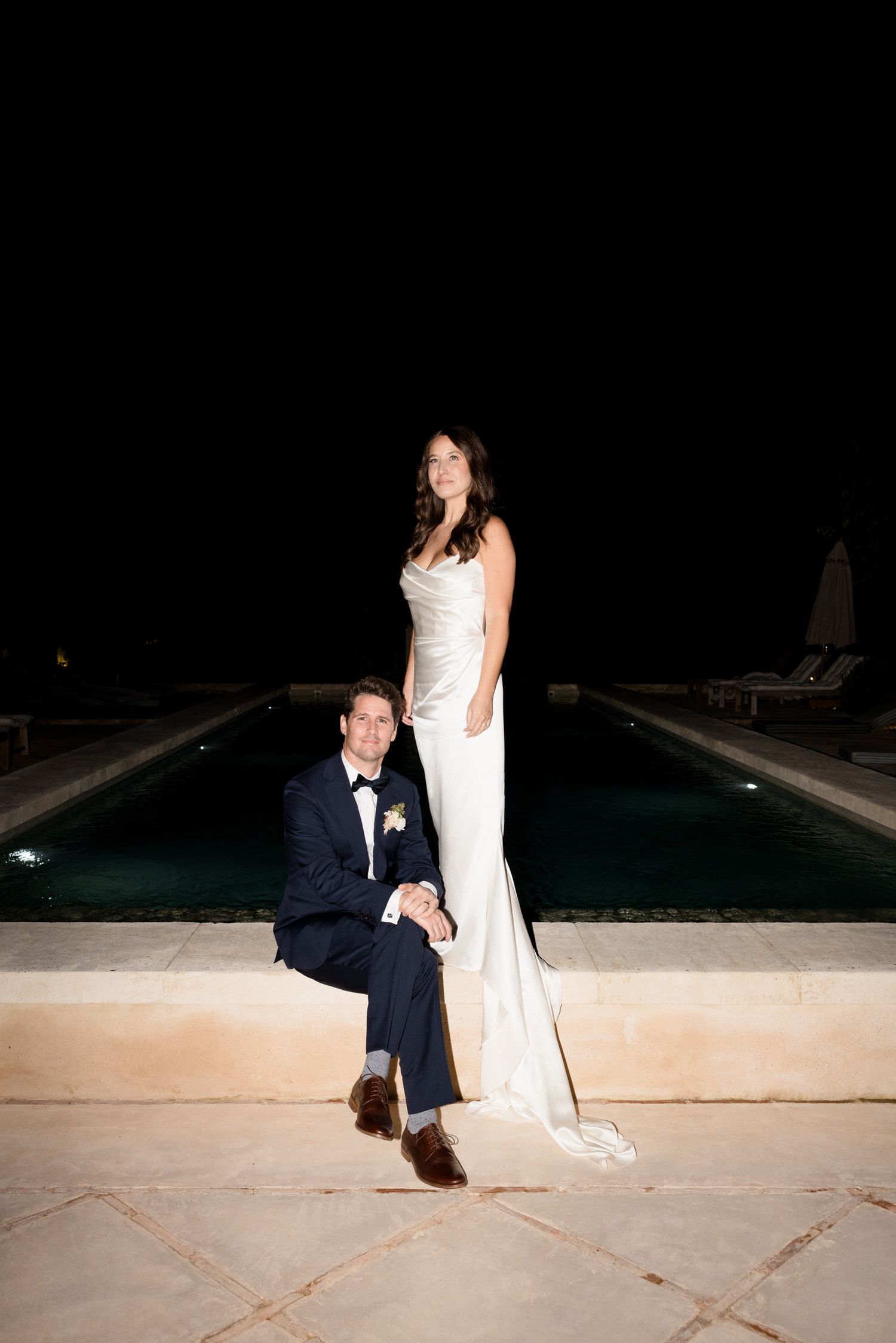 A couple in formal wedding attire sits together on stone steps at night near a pool. wedding photographer france provence