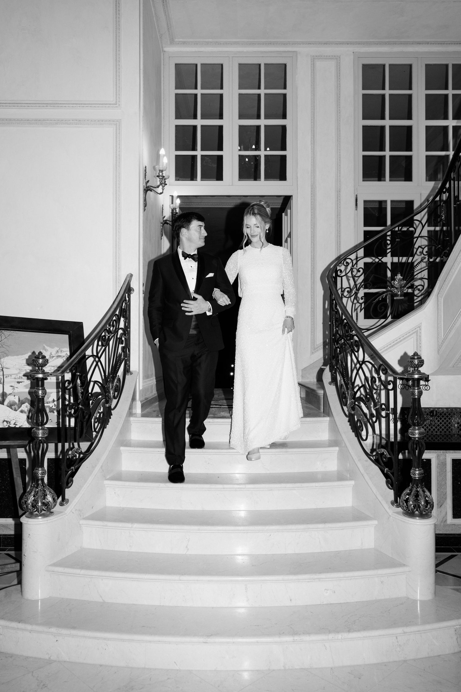 A formal couple descends an elegant white staircase with ornate iron railings in a black and white photograph. Mariage Chateau saint georges