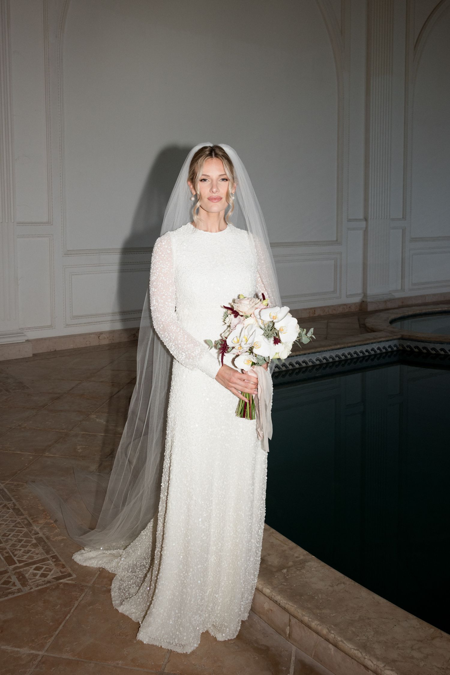 A bride in a long white lace wedding dress and veil holds a bouquet while posing in an elegant interior setting. wedding photograpapher FR