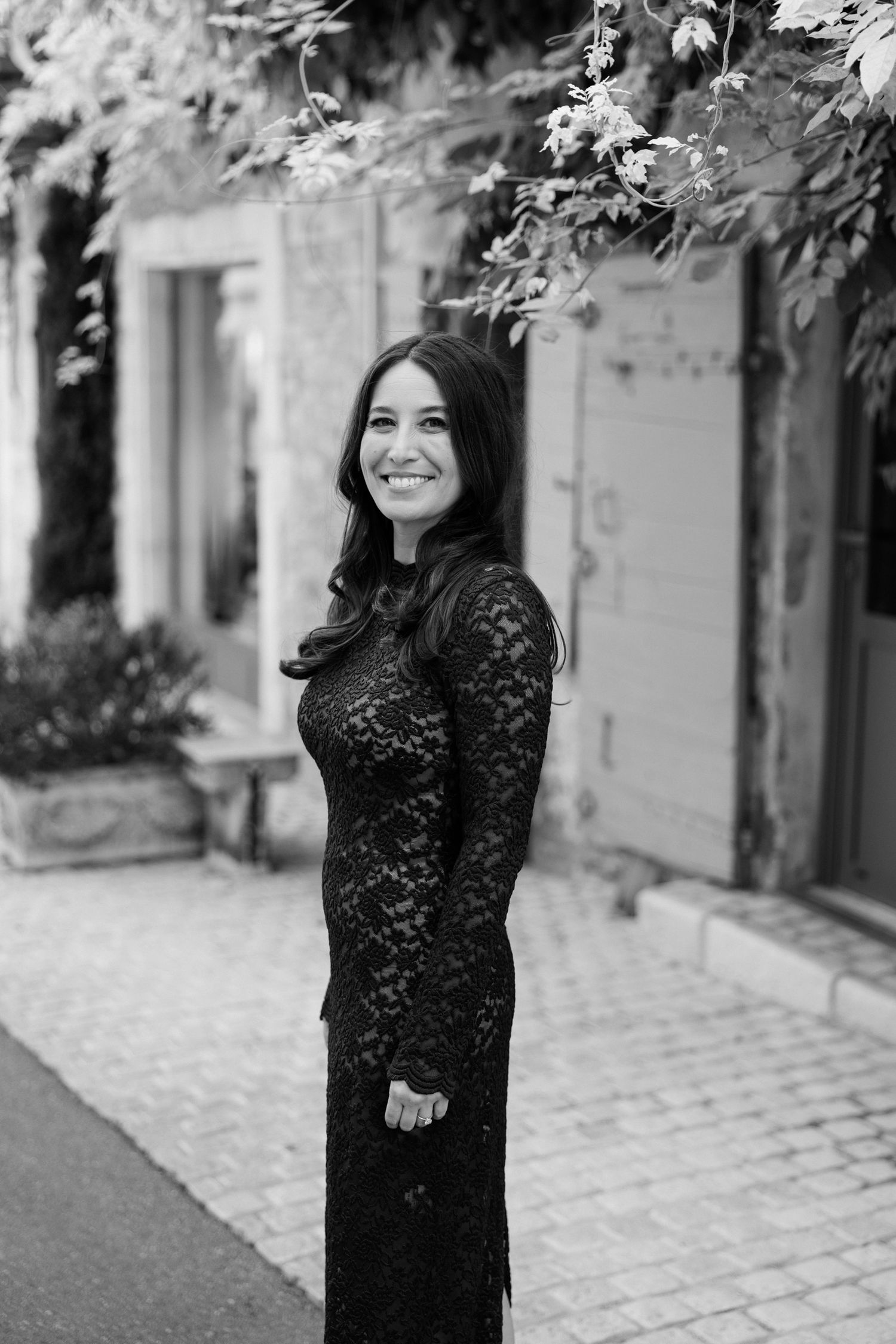 A woman in a black lace dress stands on a cobblestone path next to a building with foliage overhead. Saint Remy de Provence