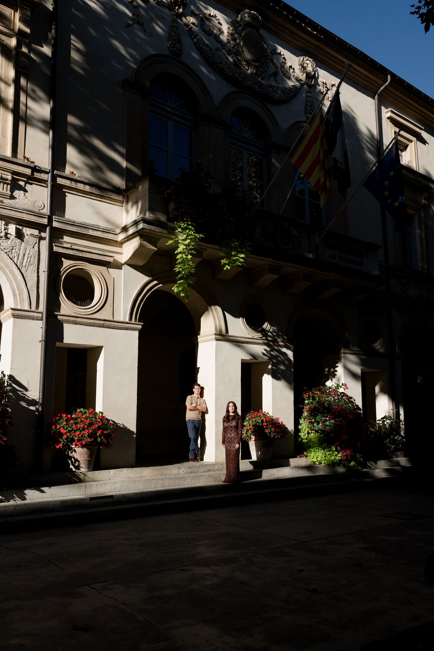 Sunlight and shadows cast dramatic patterns along a white colonnade walkway with hanging plants and architectural details.