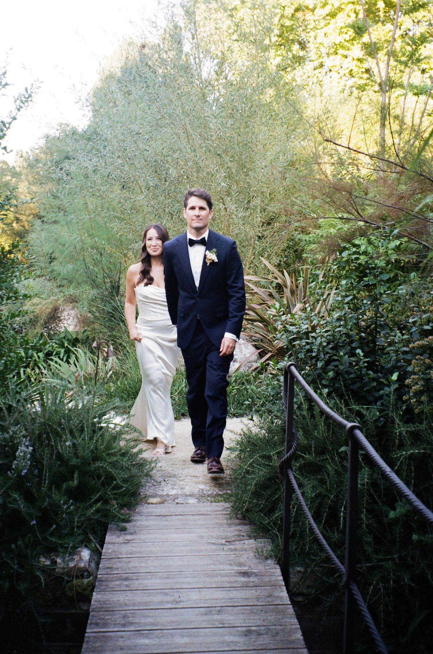 A couple in formal attire walks along a wooden boardwalk path surrounded by lush greenery in an outdoor setting.