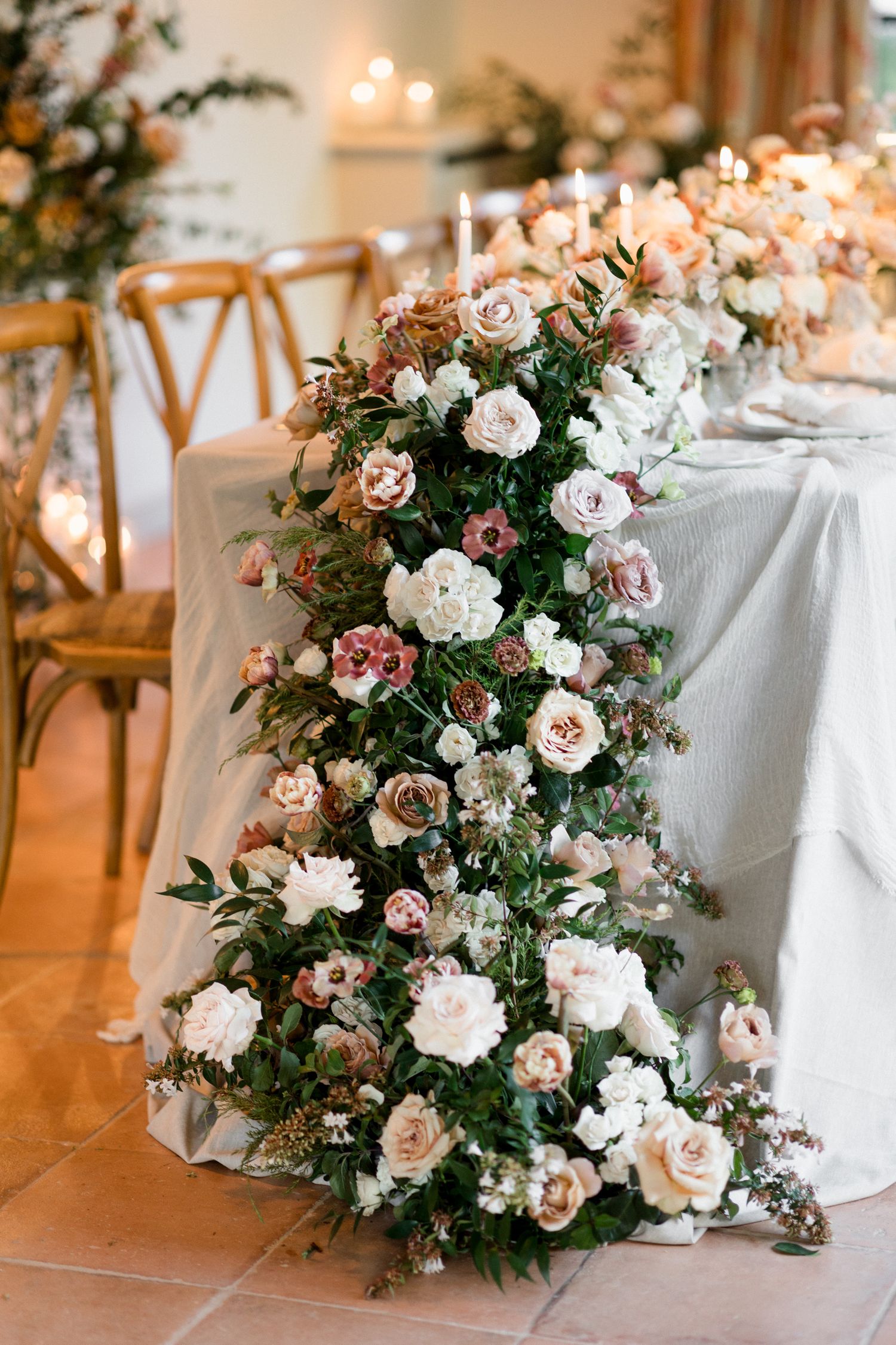 Elegant floral arrangements with white and blush roses cascade along white tablecloths at a wedding reception. wedding photographer france