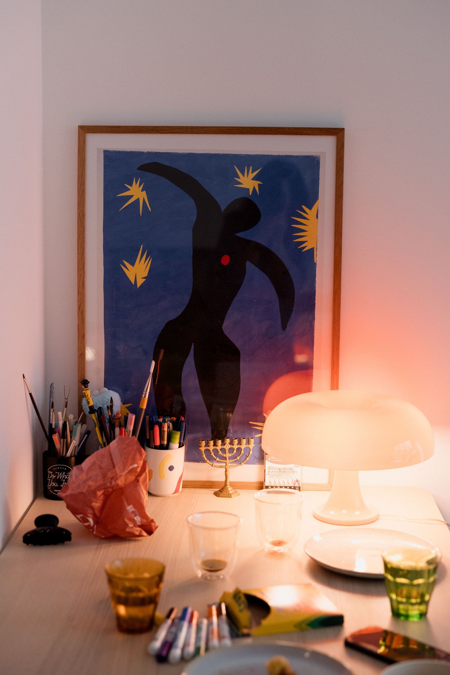 A cozy table display with a glowing mushroom lamp, menorah, art supplies, and a framed blue Matisse-inspired artwork.